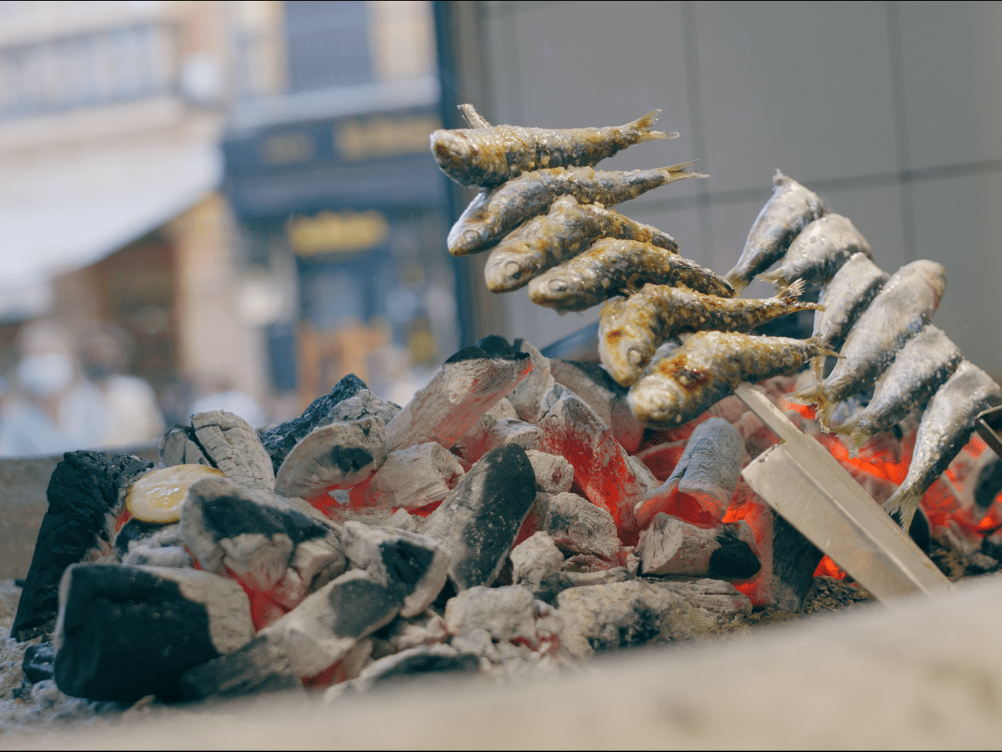 Sardines on skewers roasting over hot coals with a blurred street background.