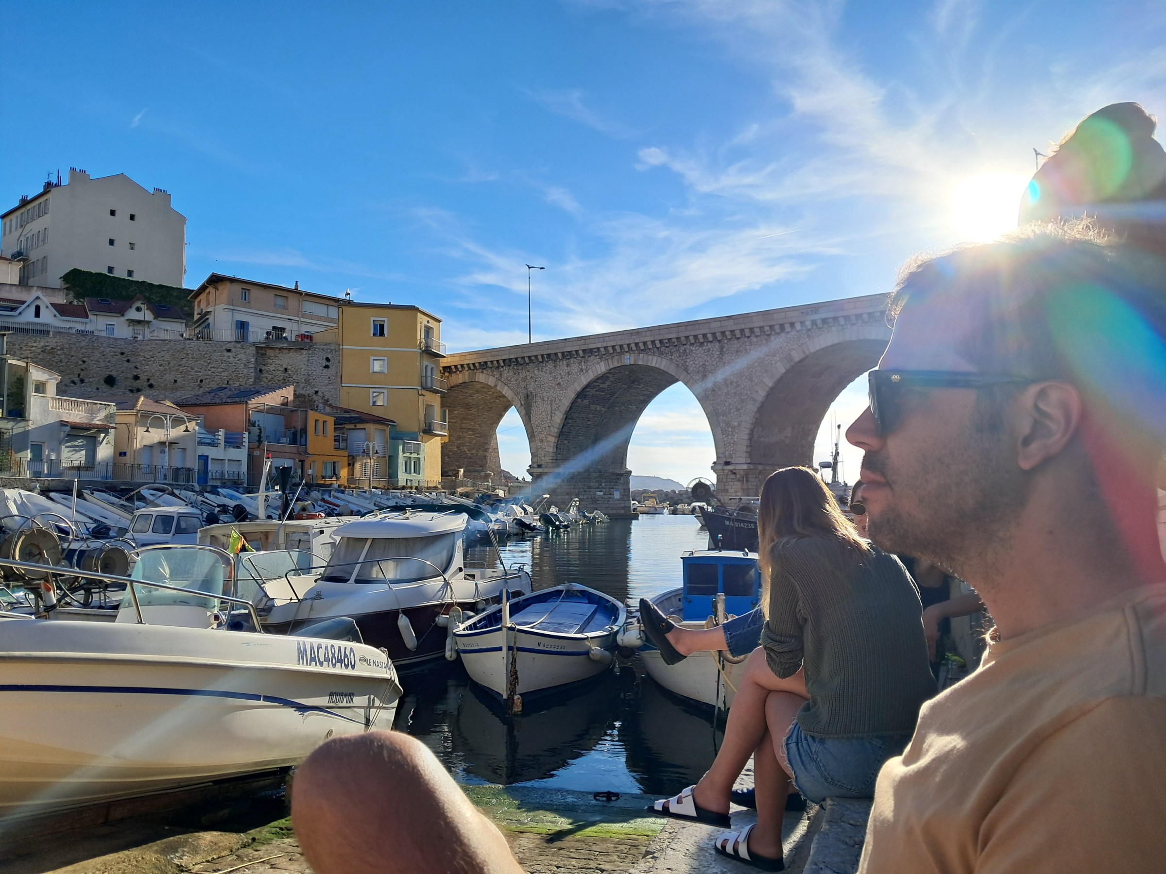 People sitting by a harbor with boats and an arched bridge under a bright sun.