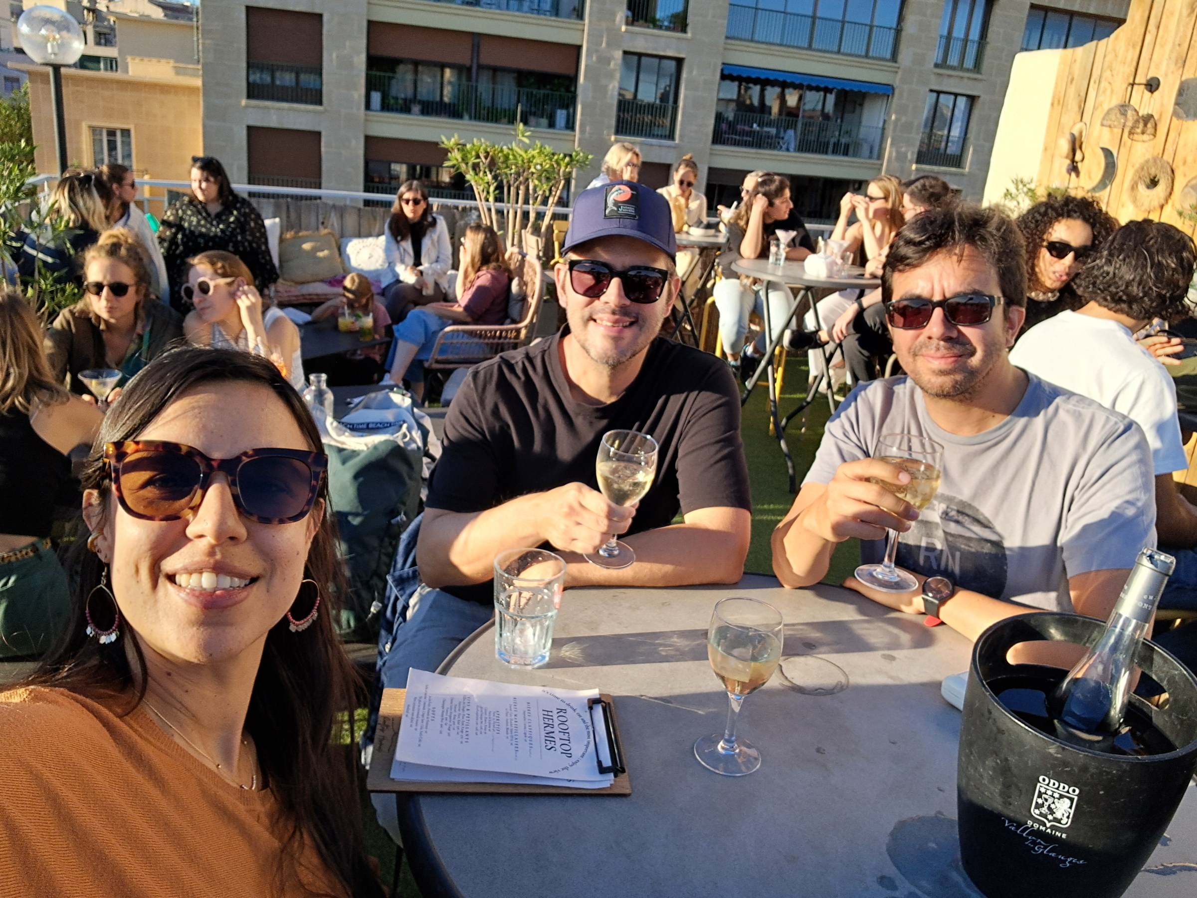 Three people smiling with drinks at an outdoor café on a sunny day.