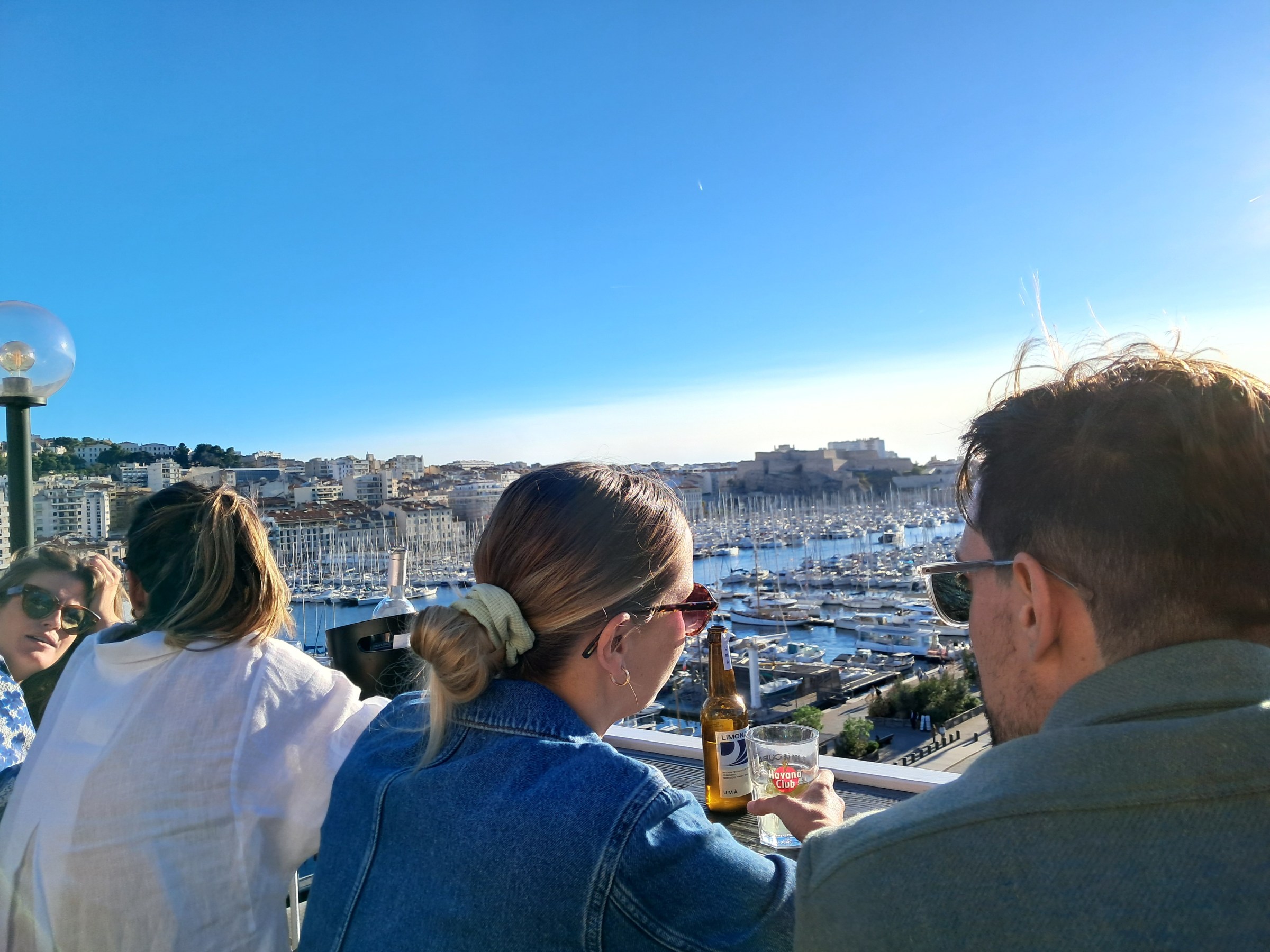 People sitting at an outdoor cafe overlooking a marina with cityscape and blue sky in the background.