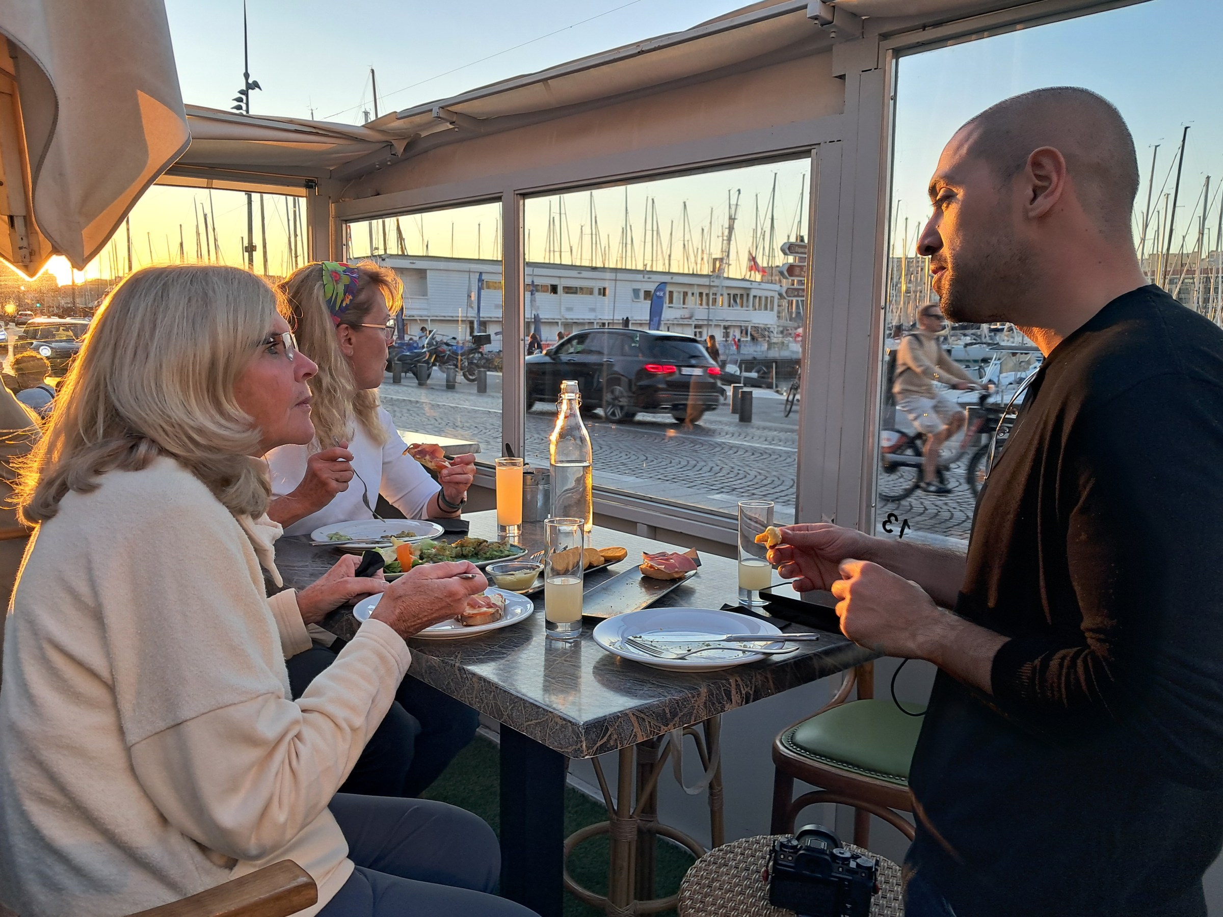 Three people dining indoors near large windows overlooking a marina at sunset.