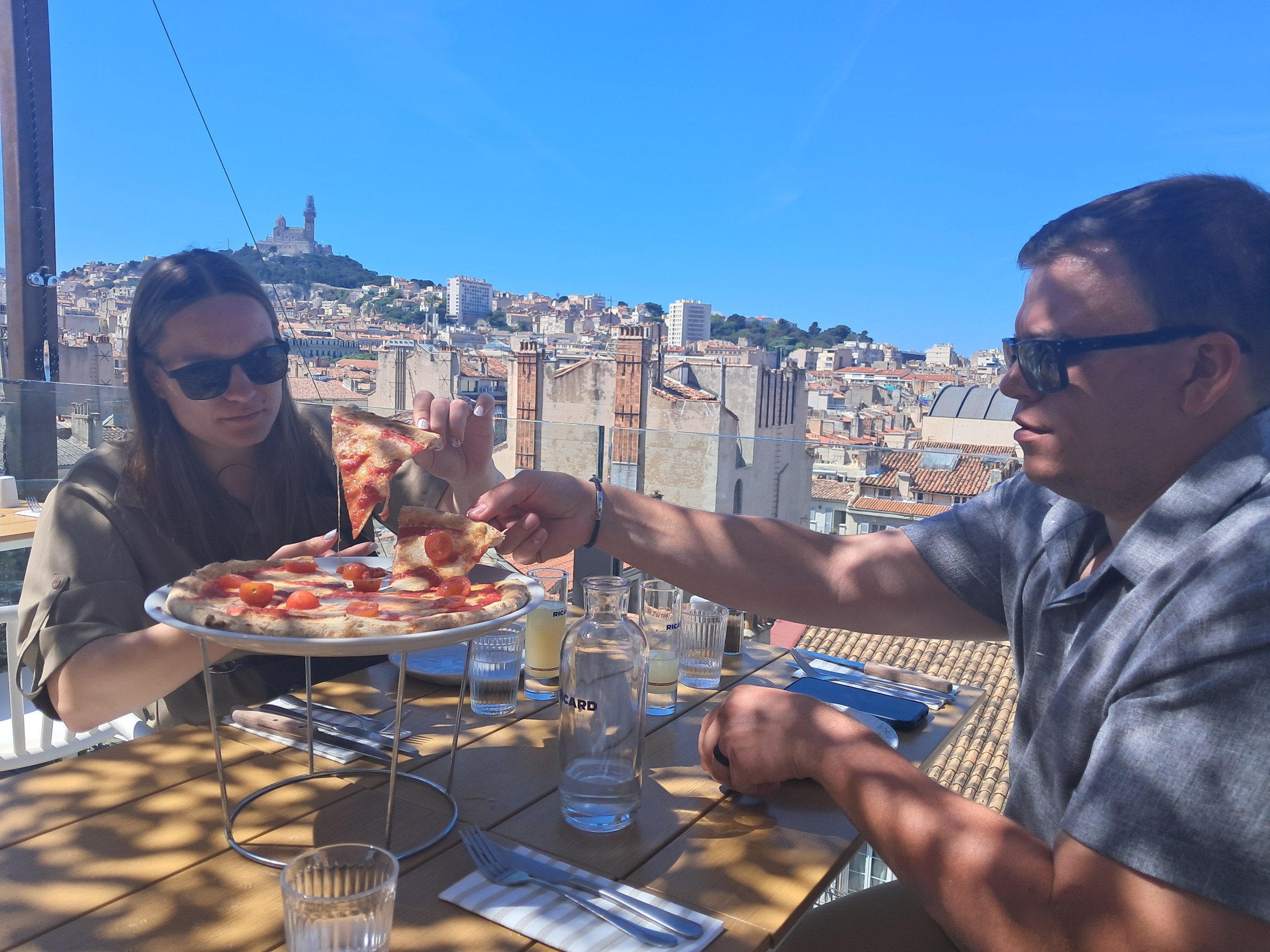 Two people share pizza at an outdoor table with a city skyline and hilltop church in the background.
