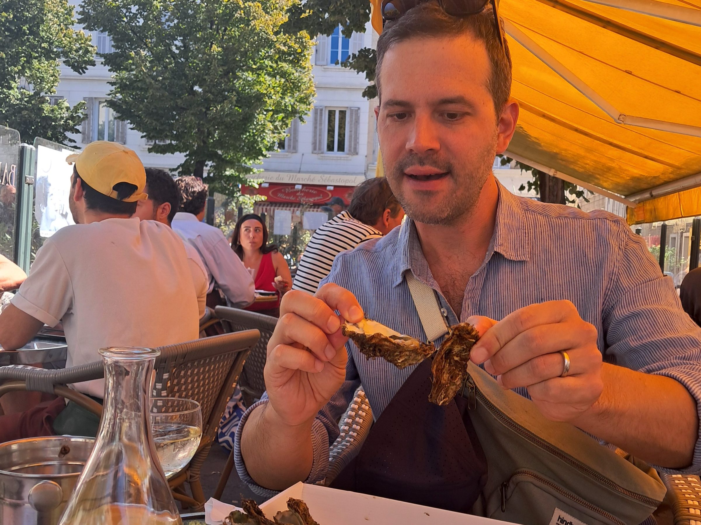 Man eating oysters at an outdoor restaurant with yellow awning and buildings in the background.