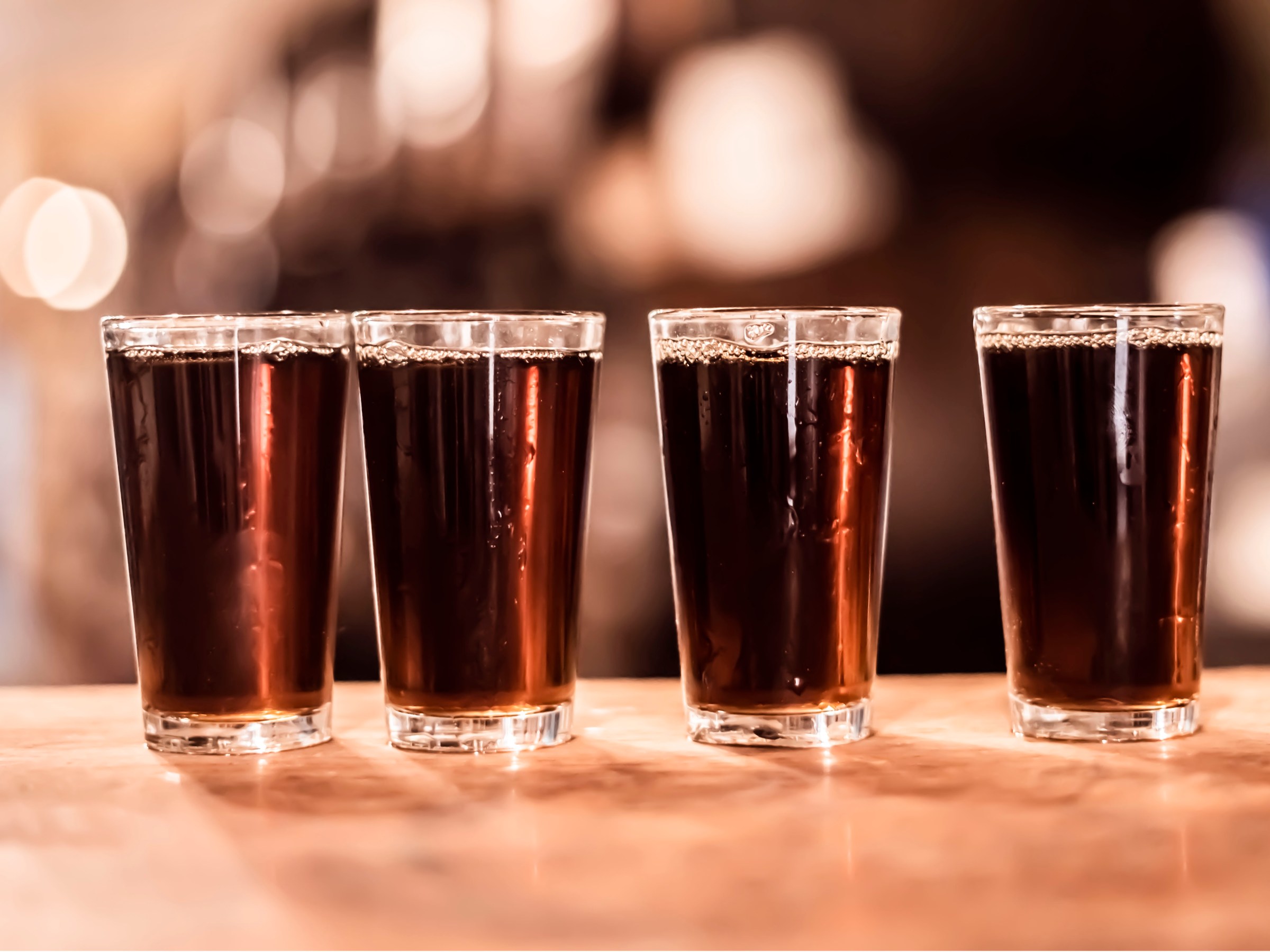 Four glasses of dark liquid on a wooden table with a blurred background.