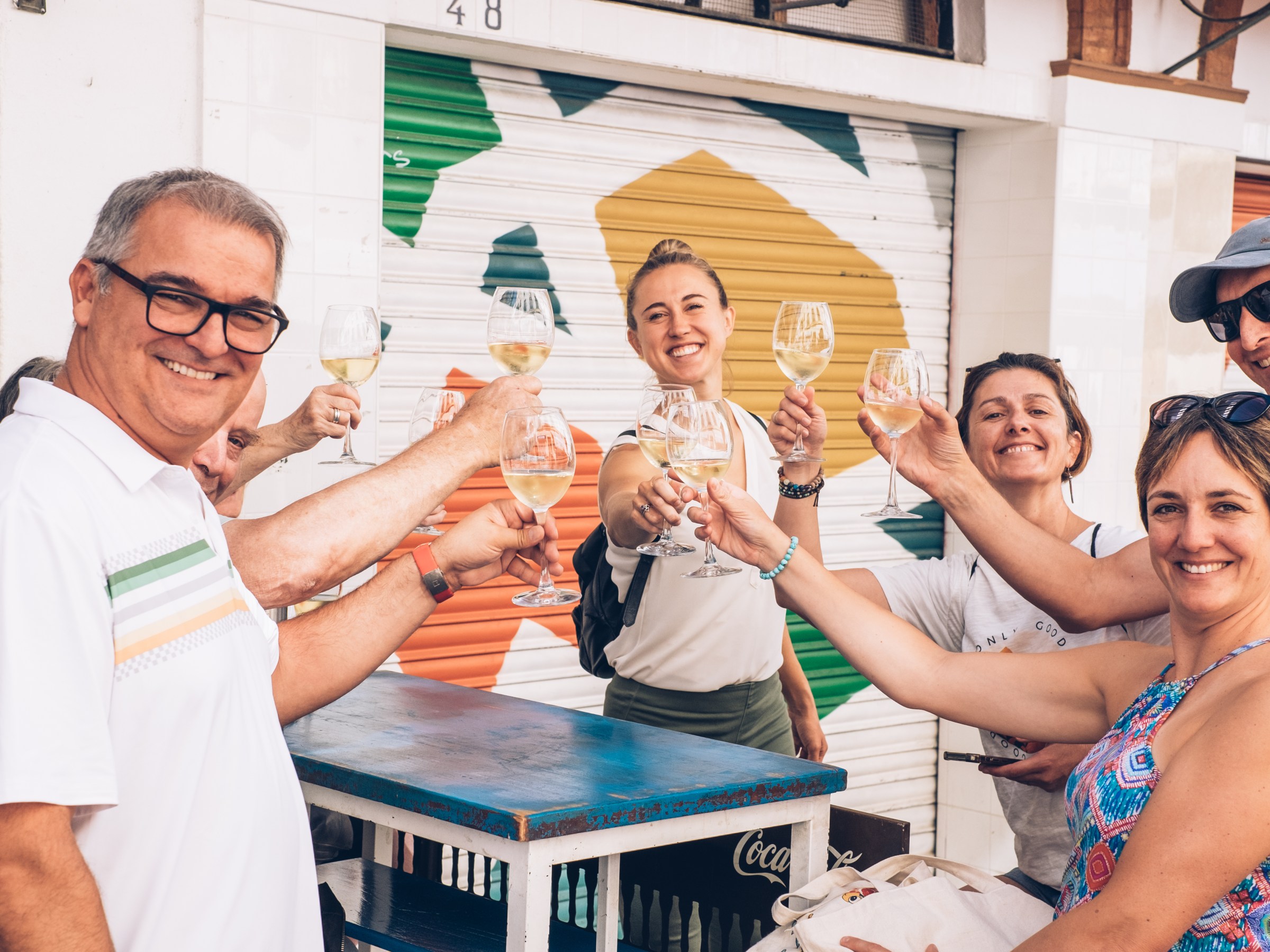 Group of people cheerfully toasting with white wine outside a colorful building.