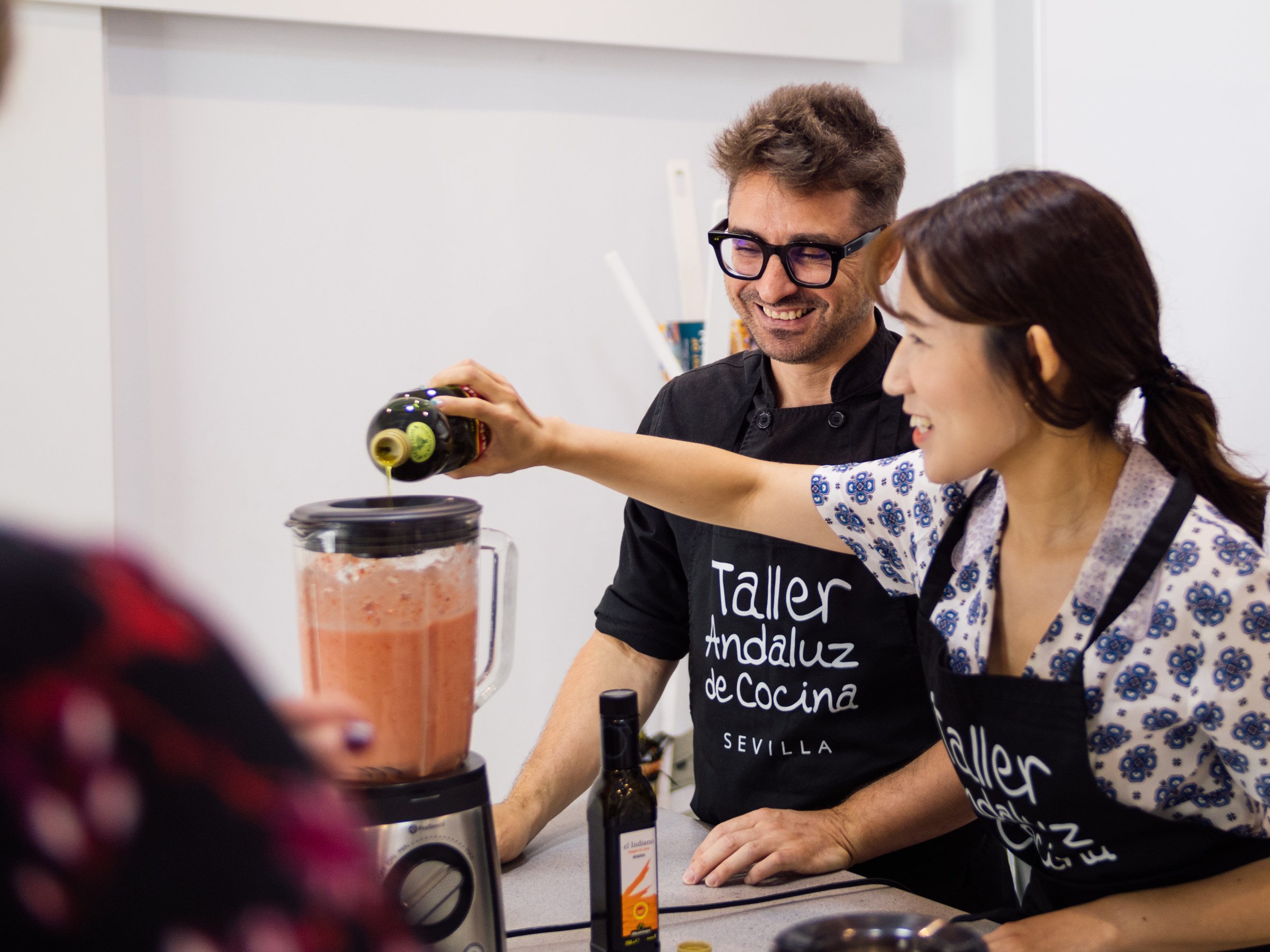 Two people in aprons making a smoothie in a blender, one pouring olive oil.