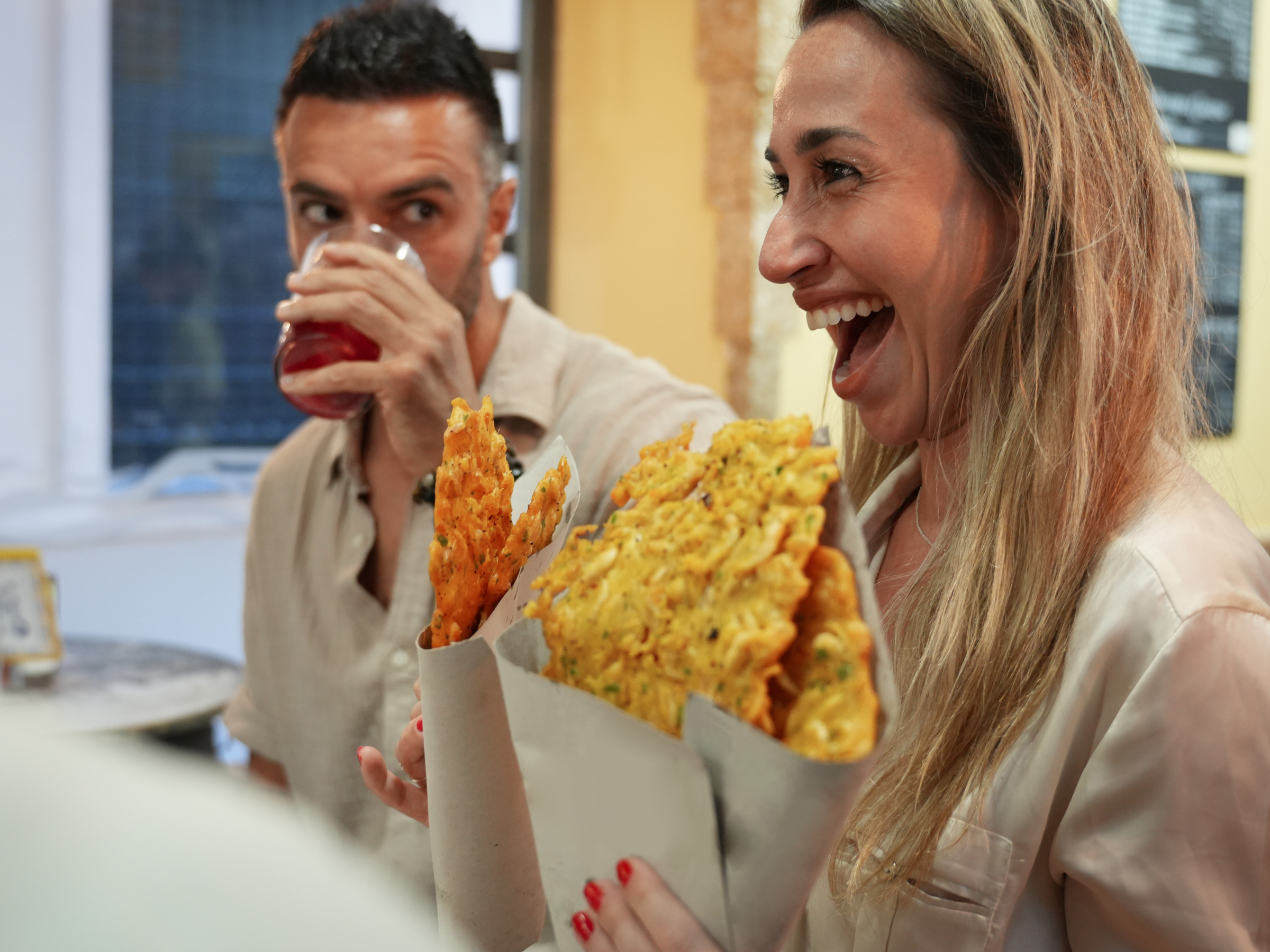 Woman smiling with fried snacks, man behind her drinks a beverage in a cafe.