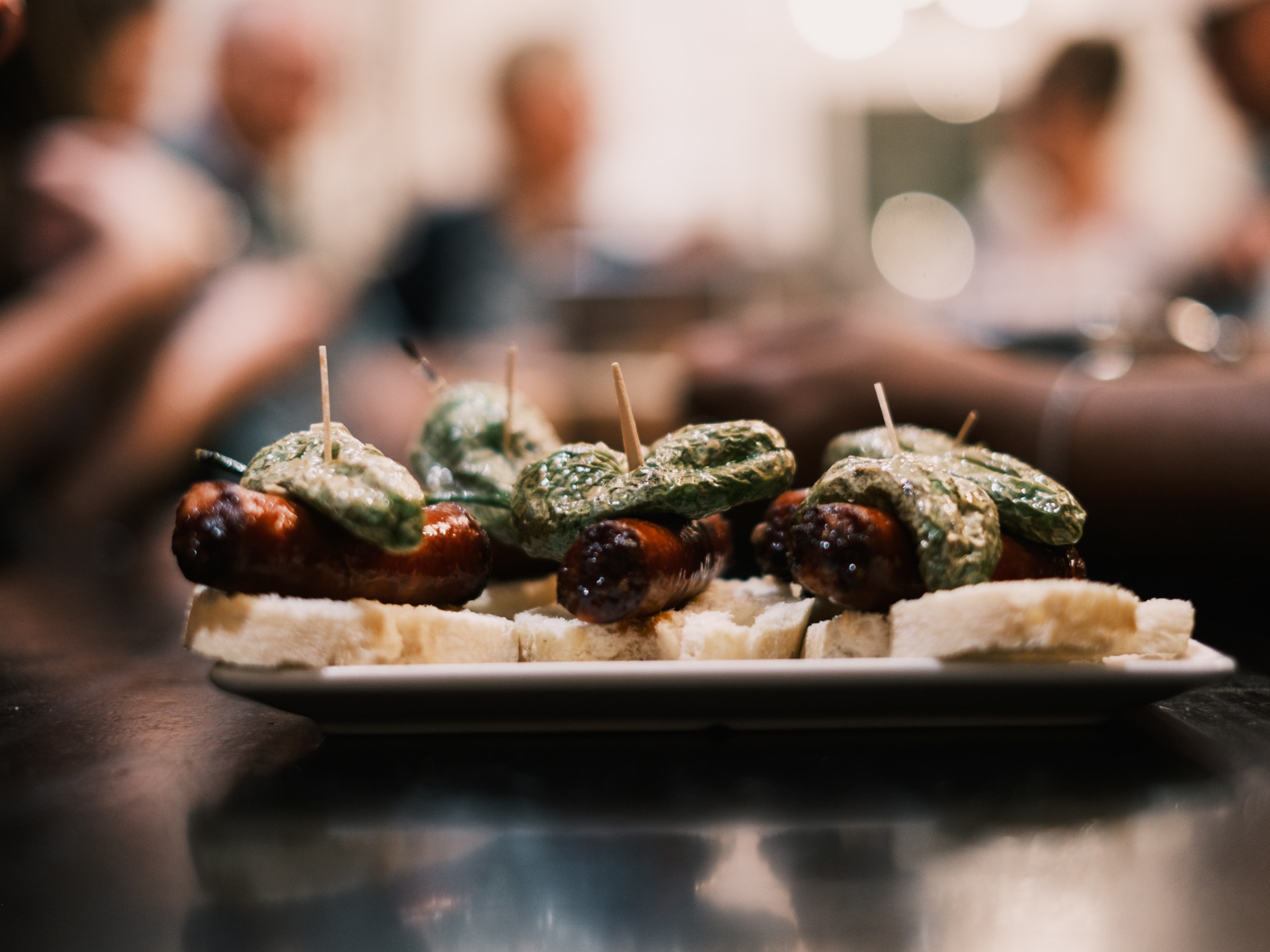 Close-up of appetizers with sausage, green peppers, and bread on a plate, blurred background.
