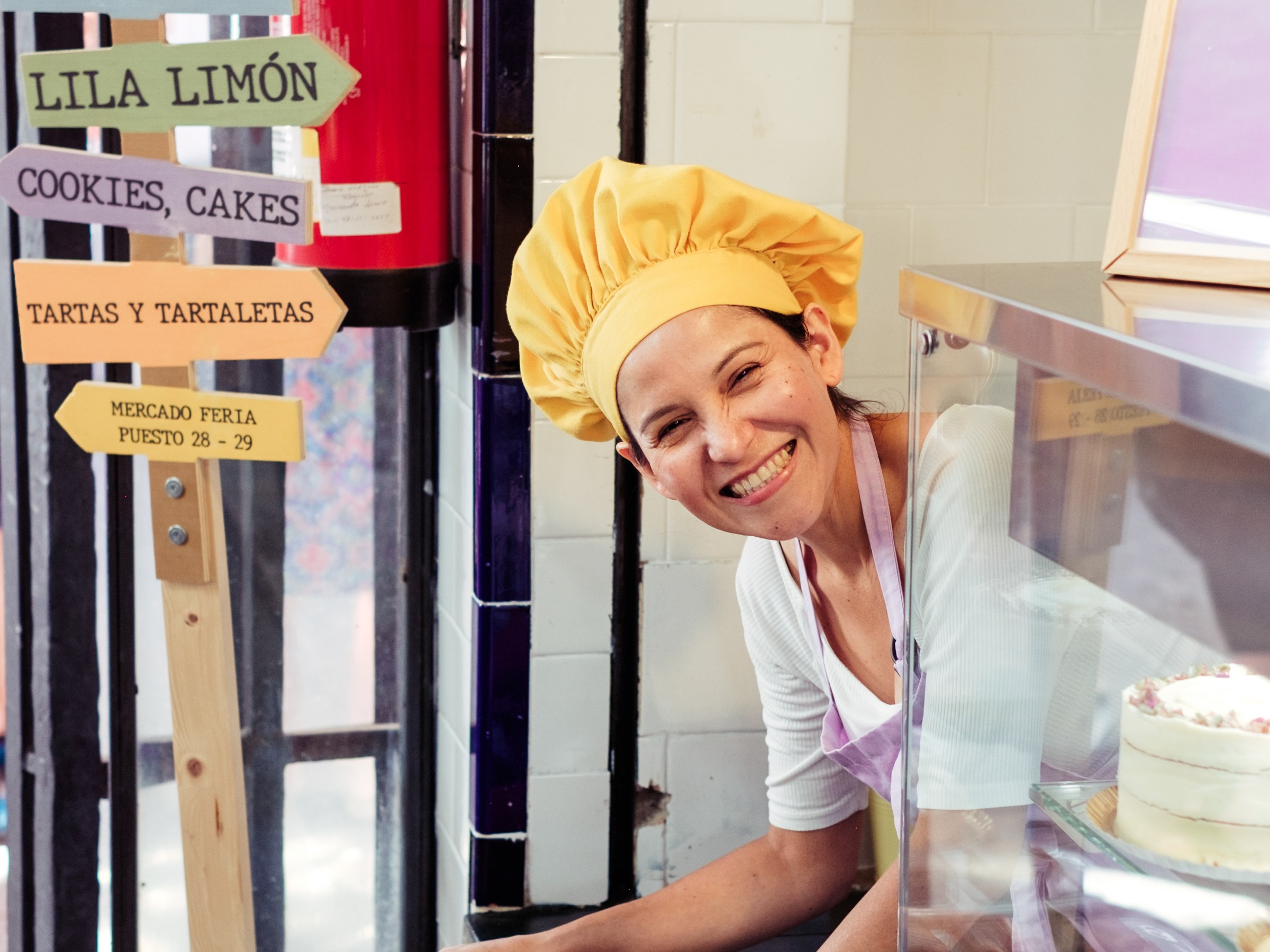 Smiling baker in a yellow hat behind a counter with colorful signs and pastries.