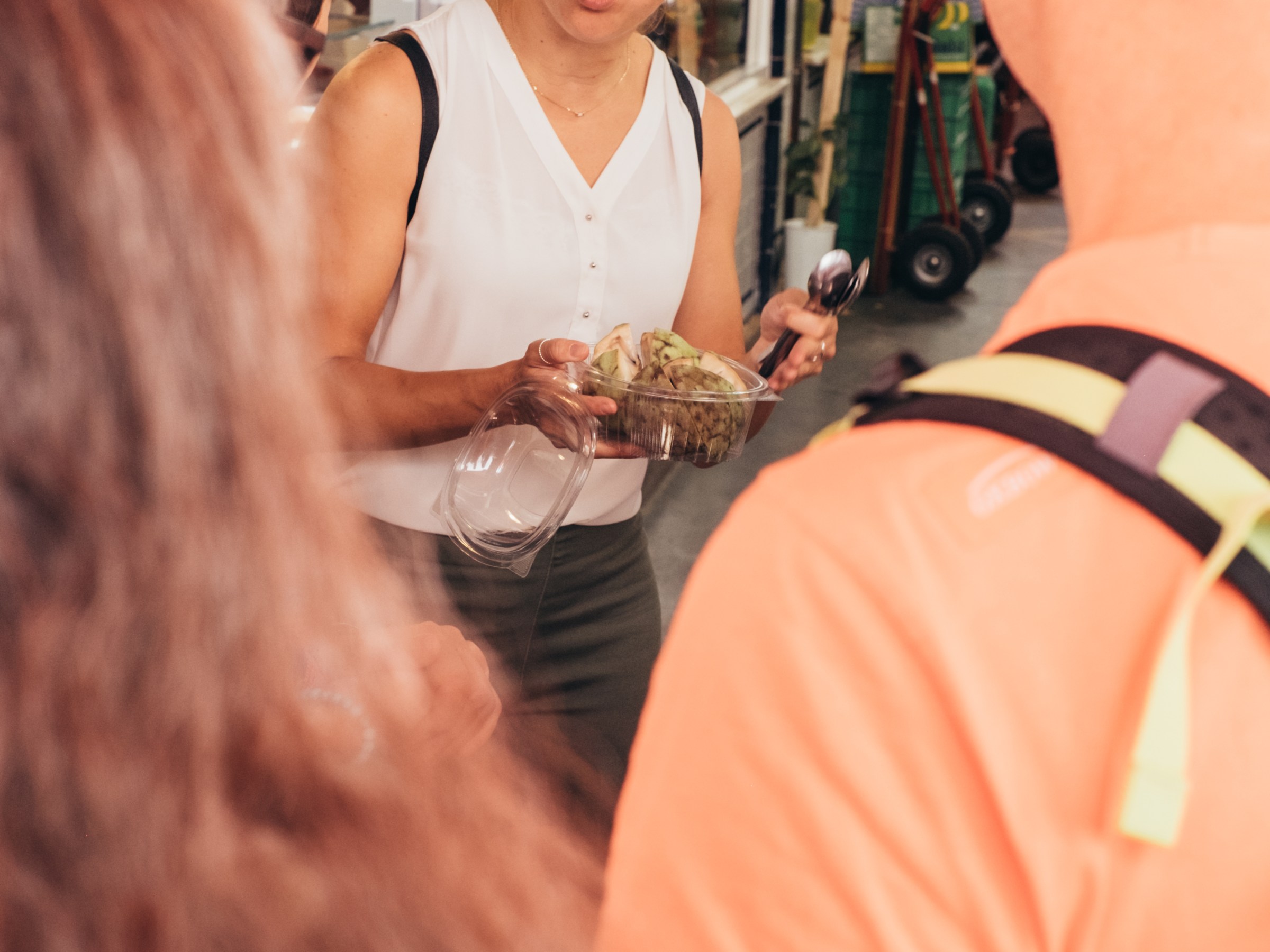 Woman with top bun talks animatedly while holding a plastic food container in a market.