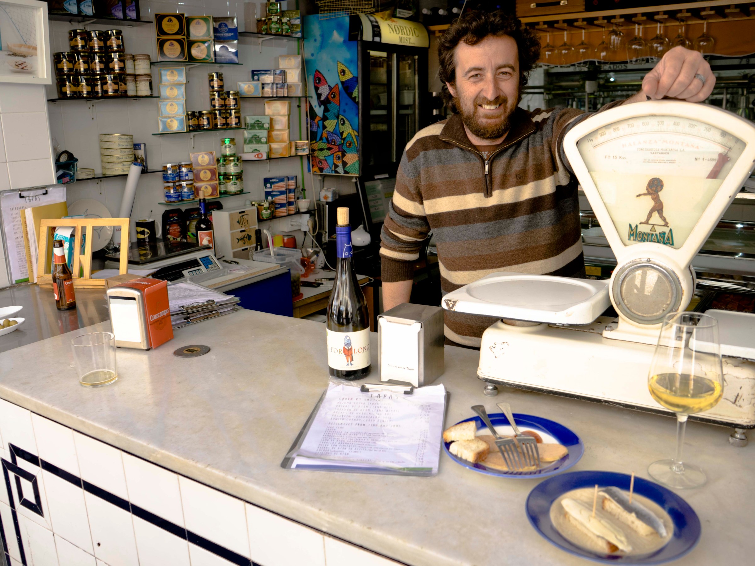 Man behind a counter with wine, food, and scale in a shop setting.