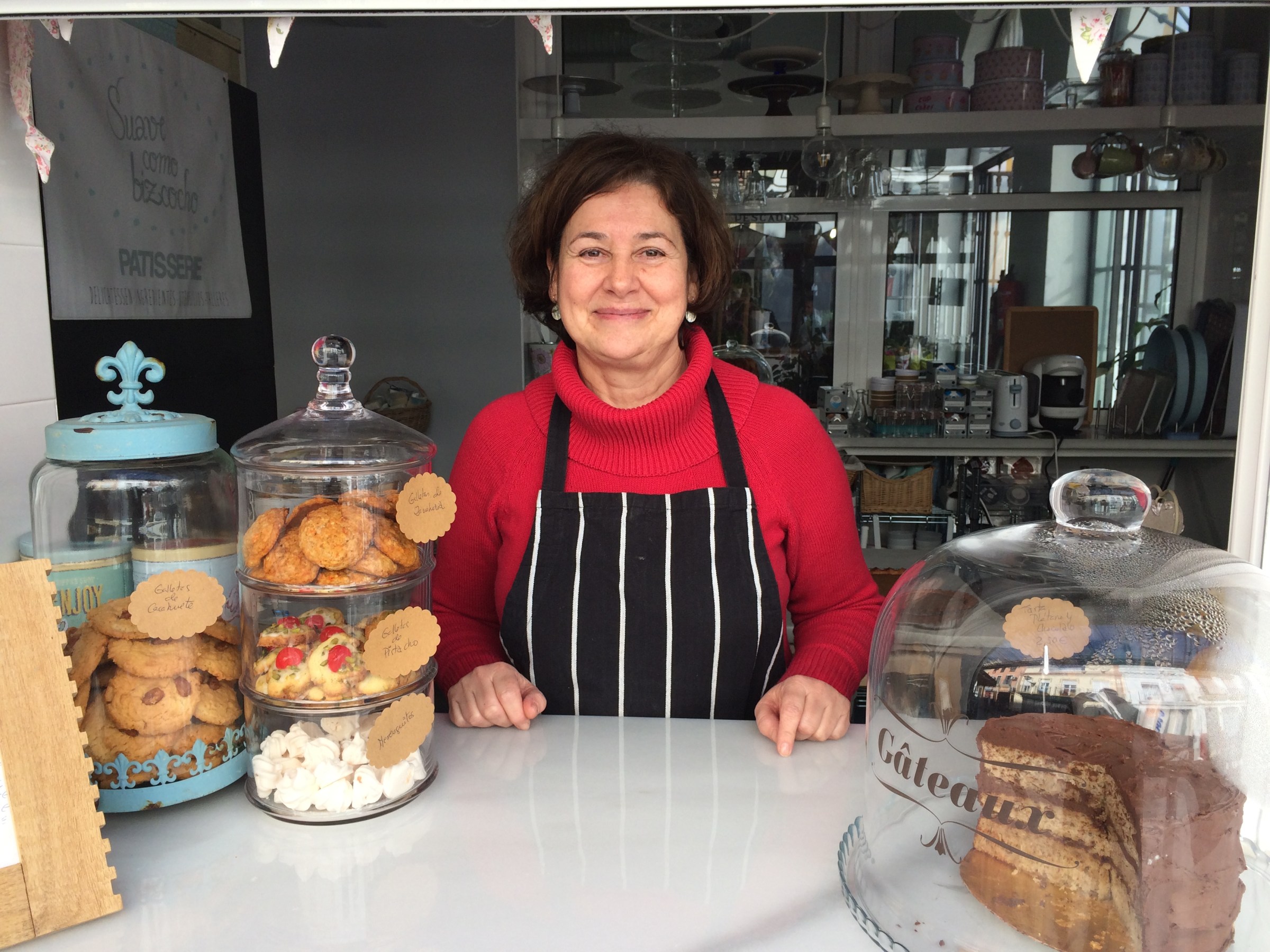 Woman in striped apron smiling behind bakery counter with assorted pastries.
