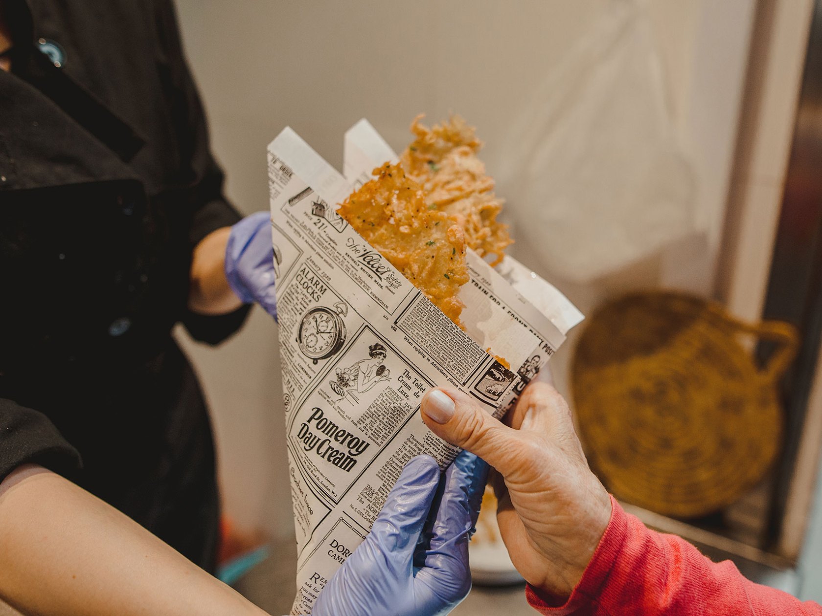 Person holding fried food in a newspaper cone from someone wearing gloves.