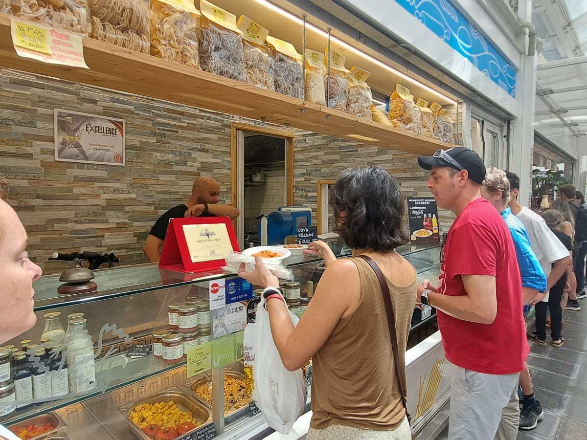 People ordering food at a market stall with pasta and sauces on display.