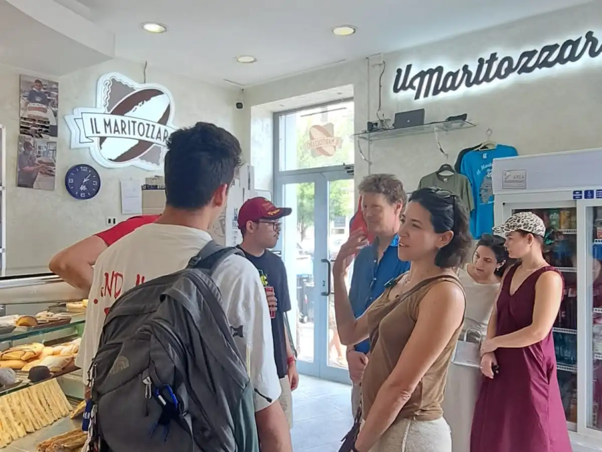 People standing inside a bakery, some looking at pastries in a display case.