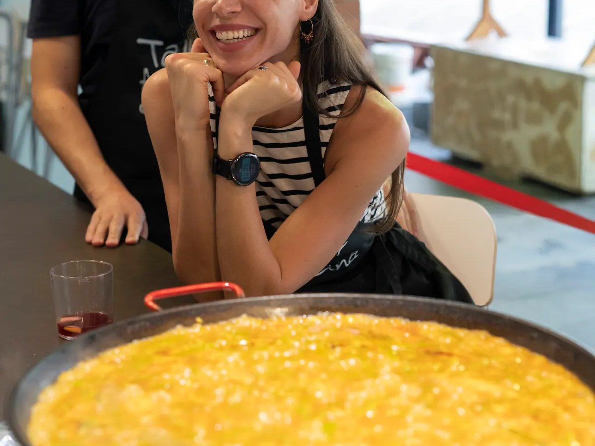 Smiling woman at a table with a large pan of bubbling food in the foreground.