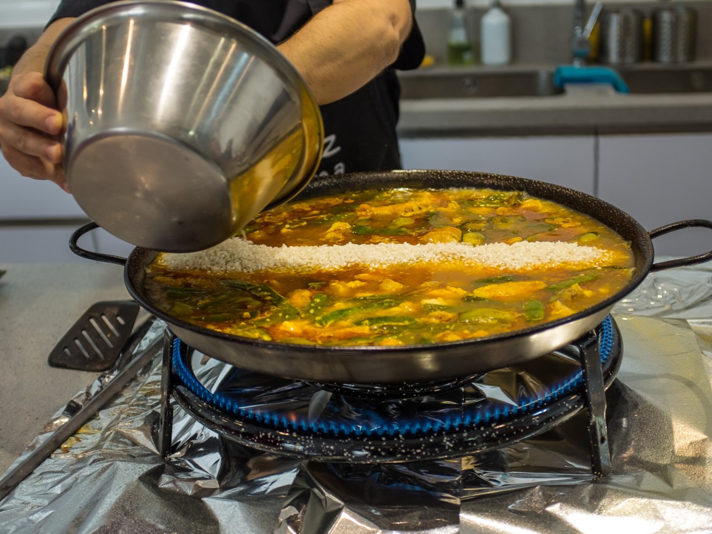 Chef pouring liquid into a large pan of yellow paella on a stove.