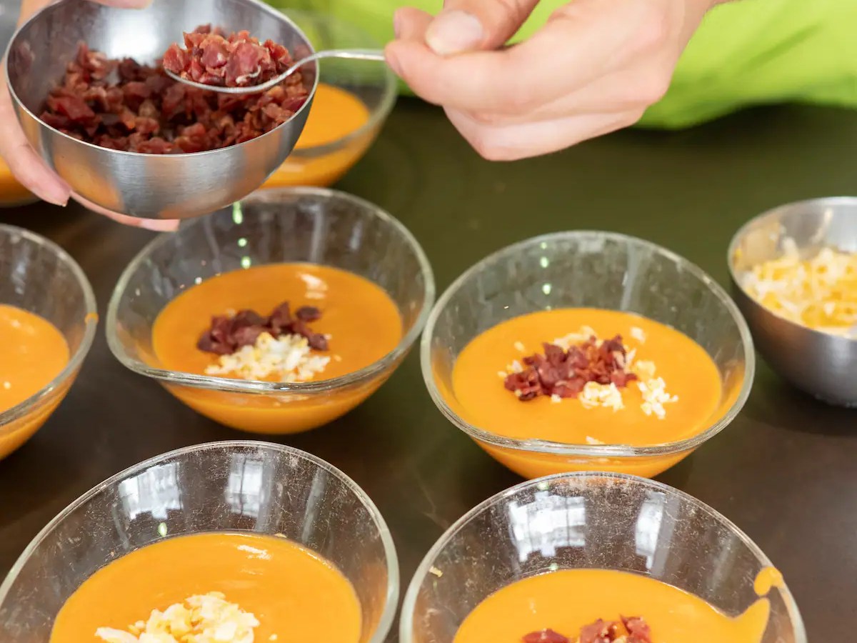 Person garnishing bowls of soup with chopped toppings from a metal bowl.