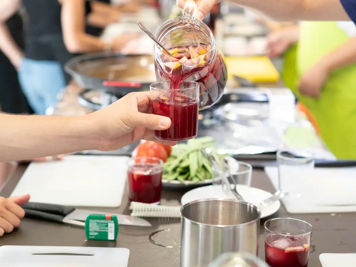 People pour red juice from a pitcher into a glass at a table with food items.