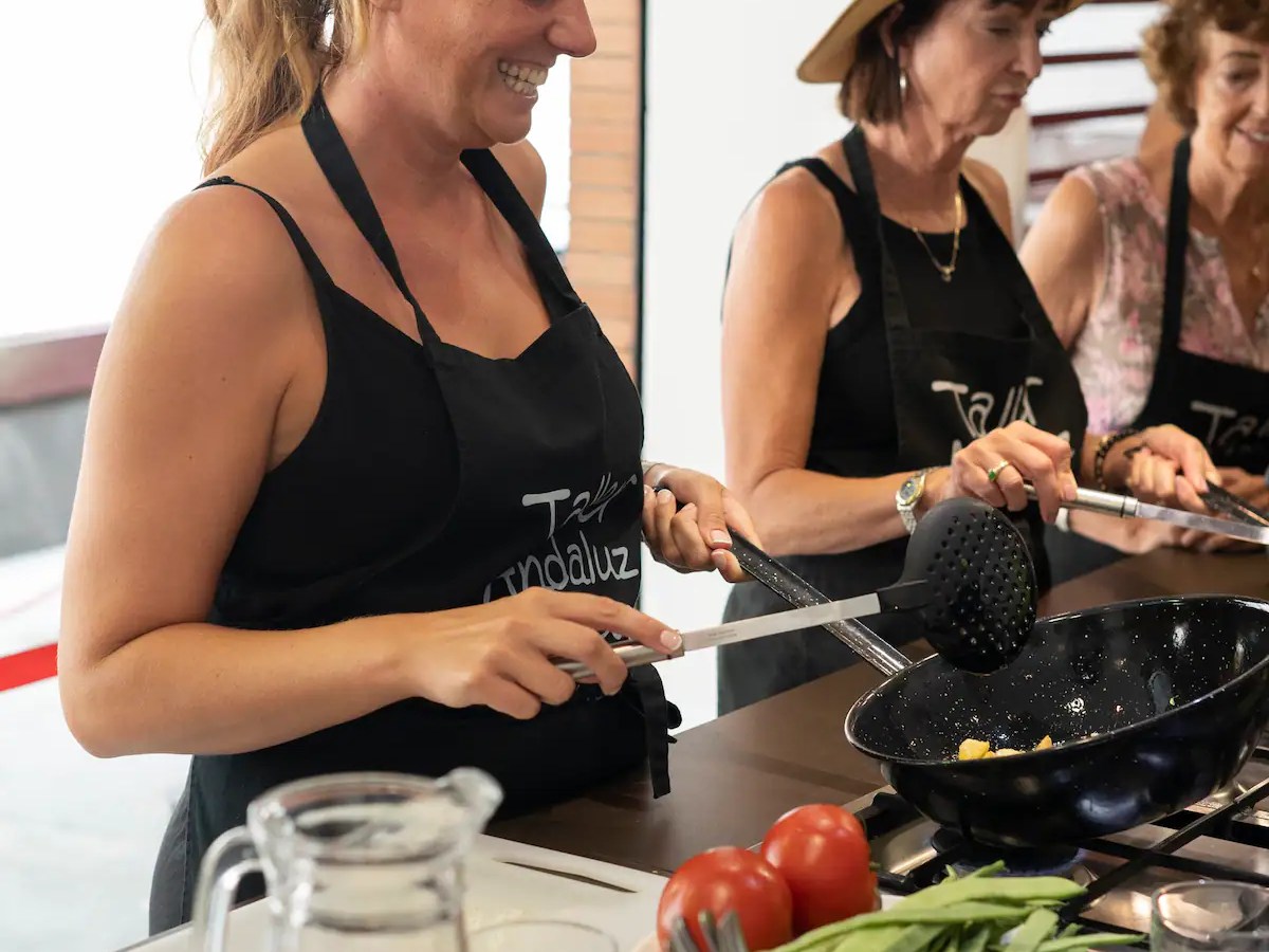 Women in a cooking class, smiling, wearing aprons, using kitchen utensils on a stove with vegetables nearby.