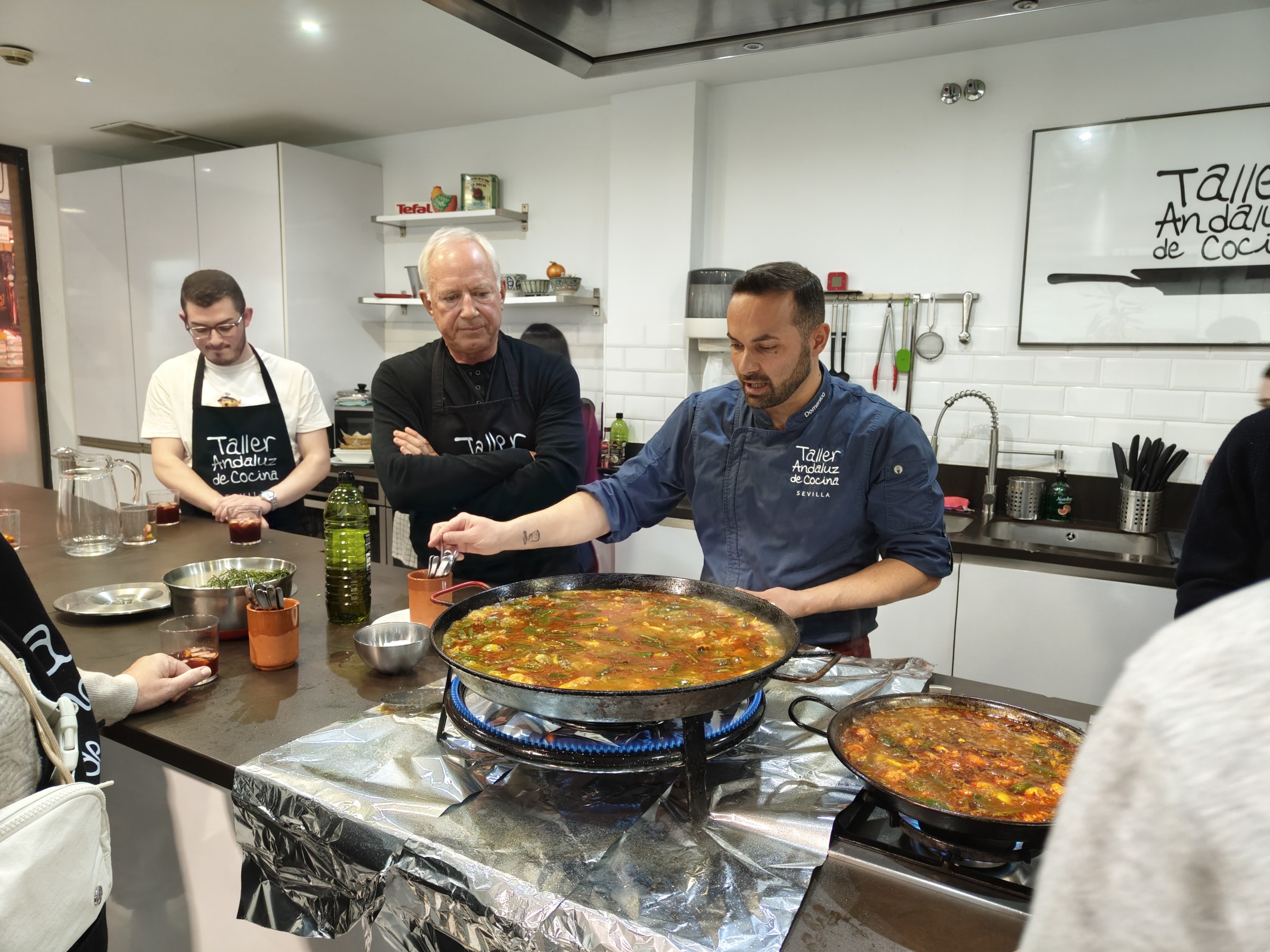 Chef demonstrating cooking technique to class around large pan in kitchen.