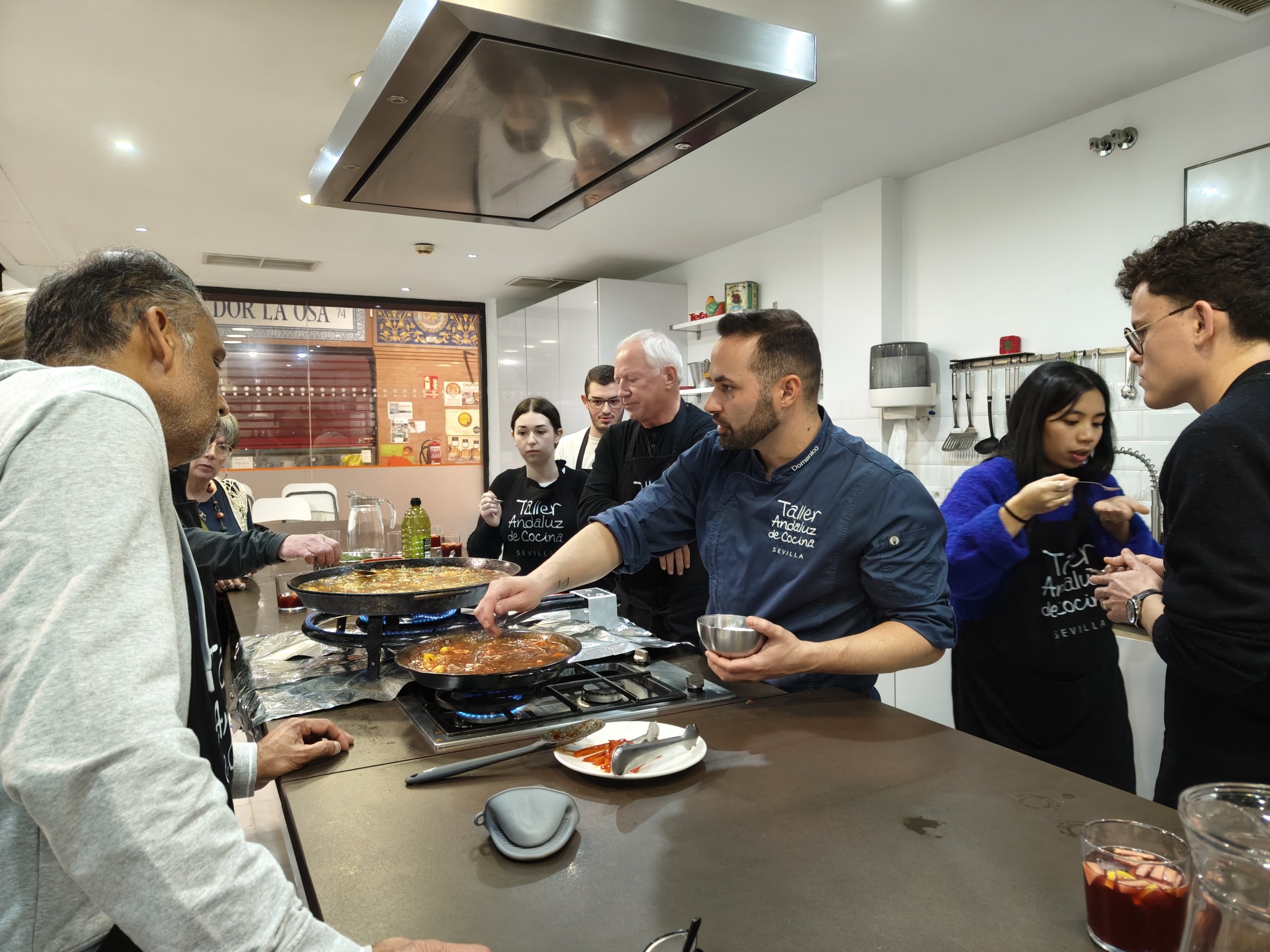 People gather around a chef cooking paella in a kitchen workshop setting.
