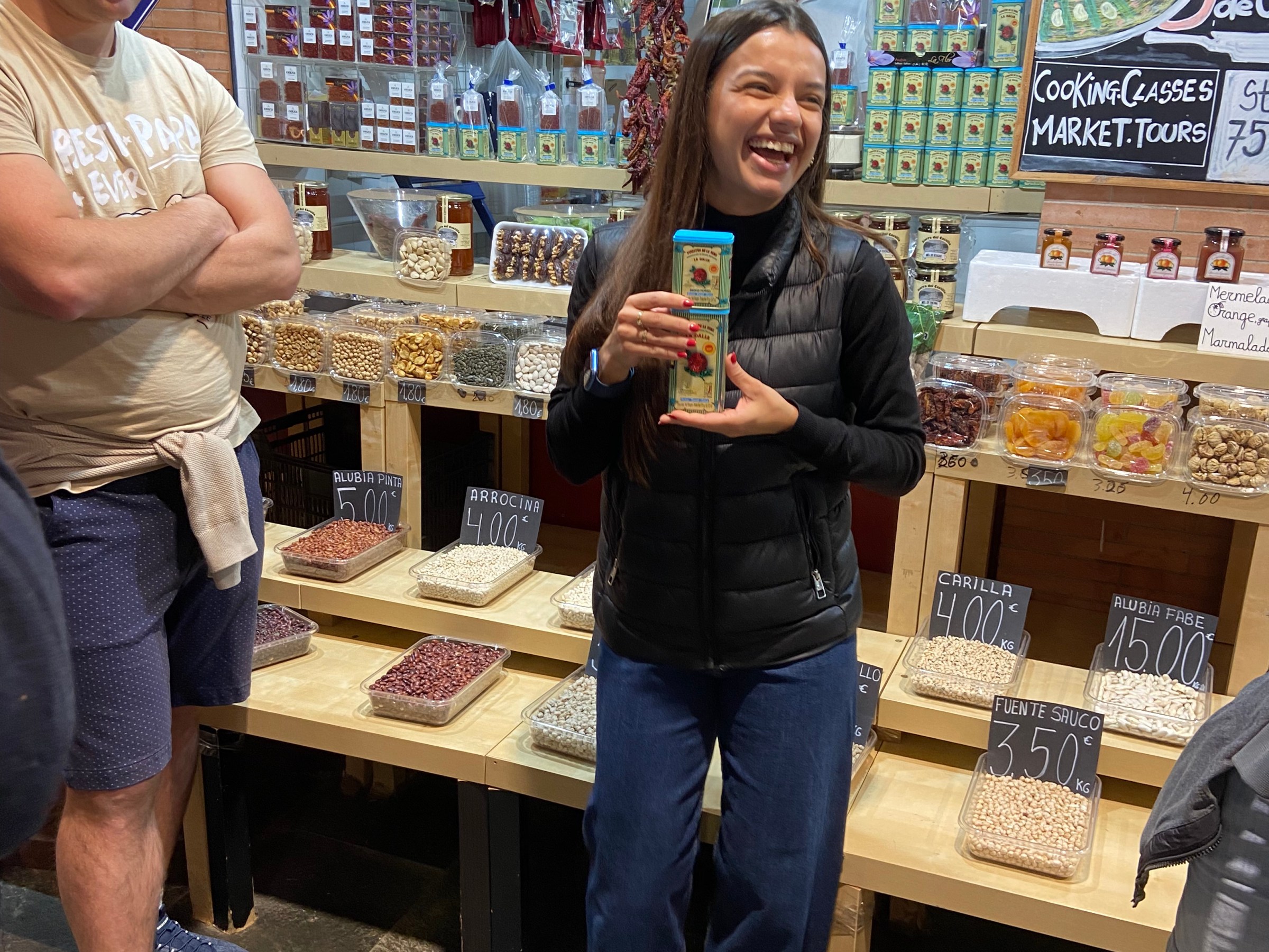Smiling woman holding a product in a busy market stall with shelves of spices and legumes.