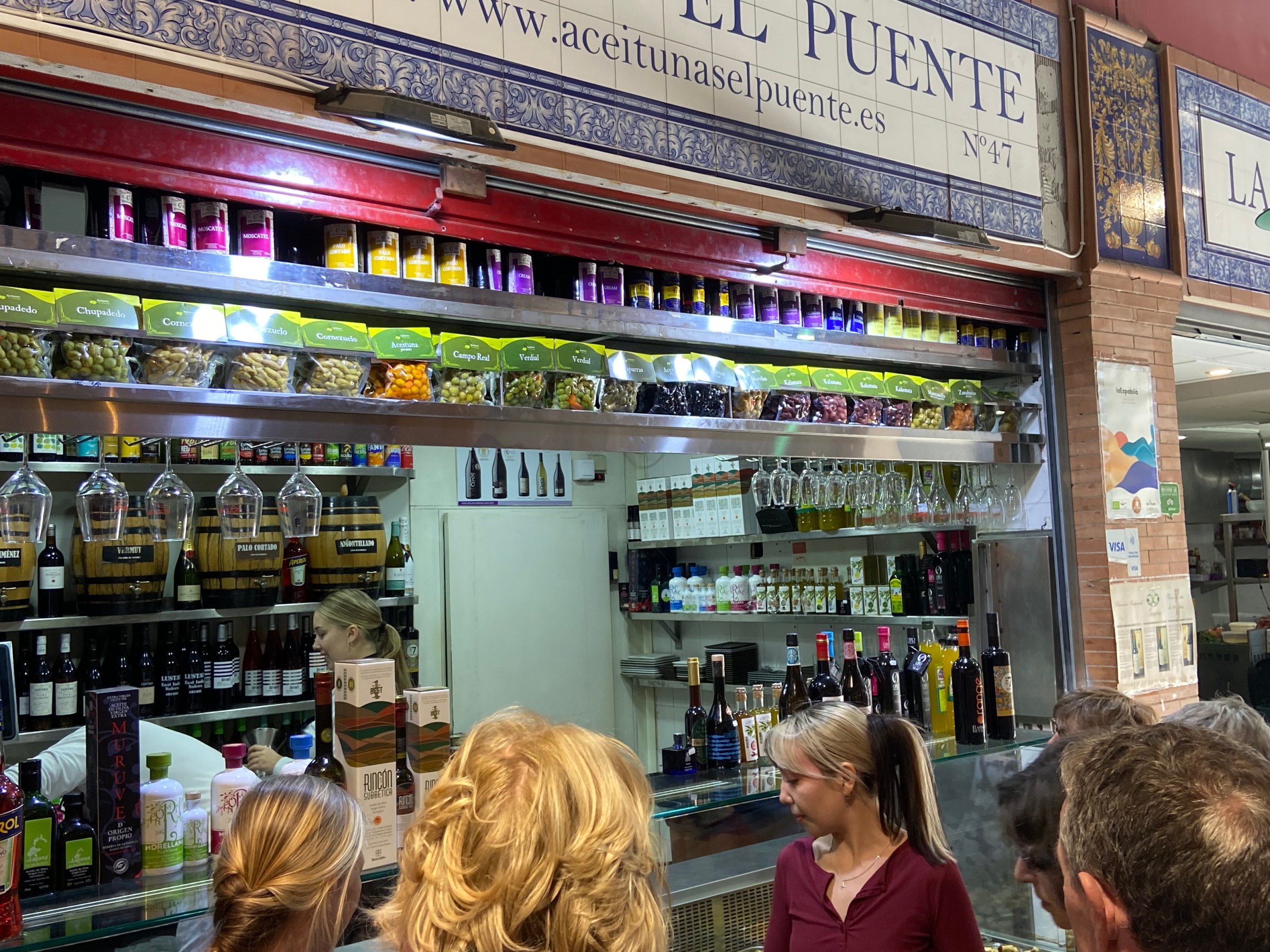 People standing at an olive and wine shop counter named Aceitunas El Puente in a market.