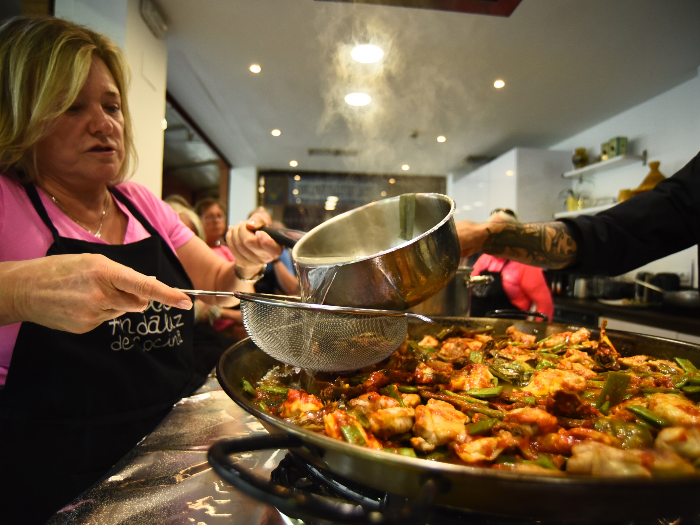 Woman in apron cooking paella, pouring liquid into a large pan with a strainer.