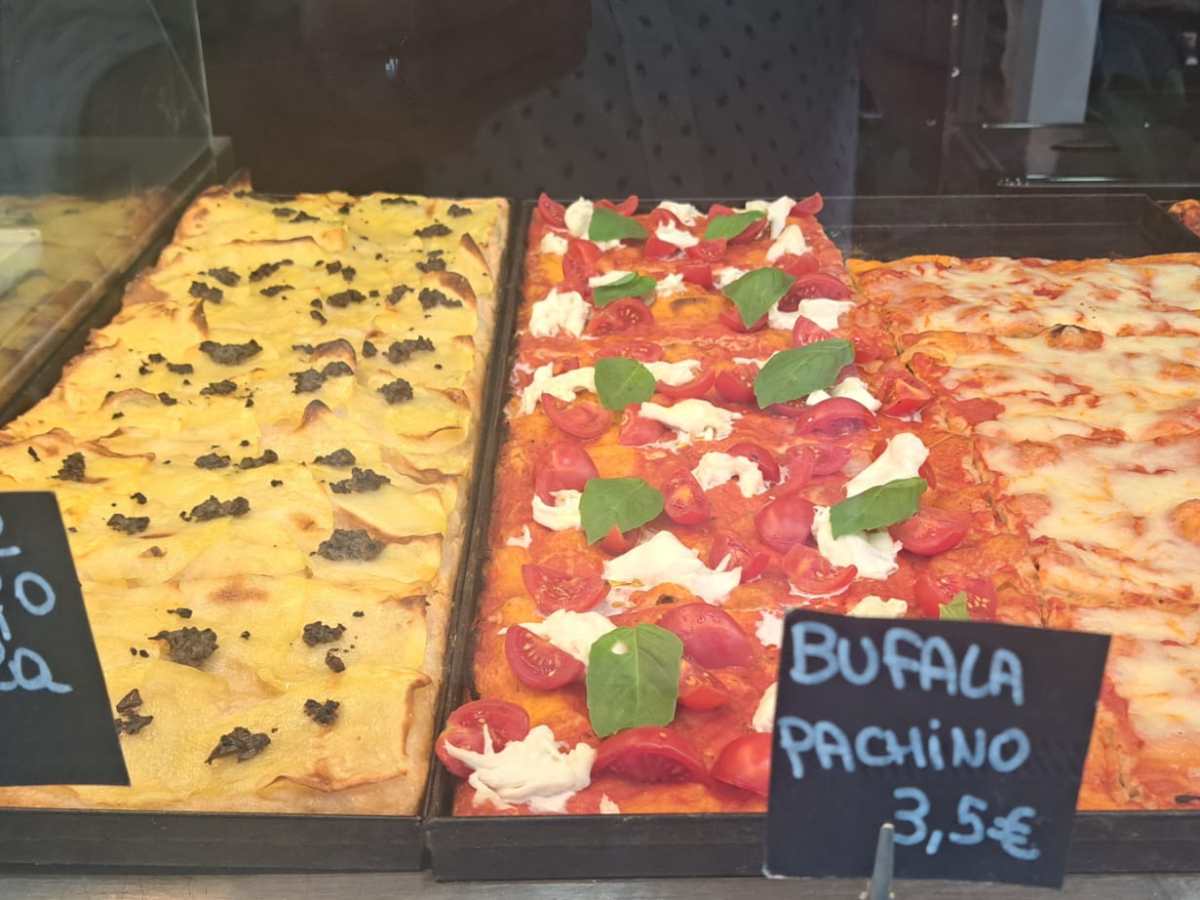 Three trays of assorted pizza slices with handwritten price signs in a display case.