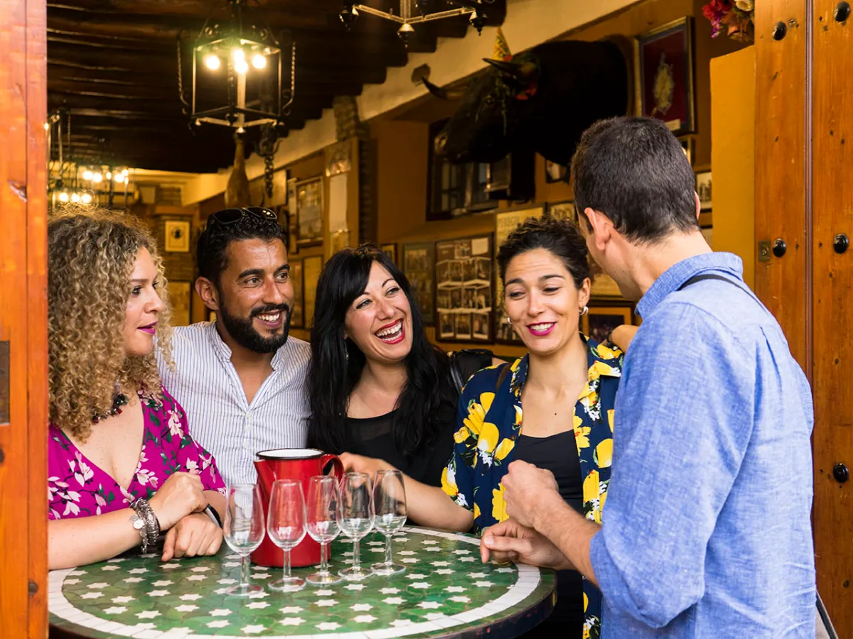 Group of five people laughing around a round table in a cozy bar setting.