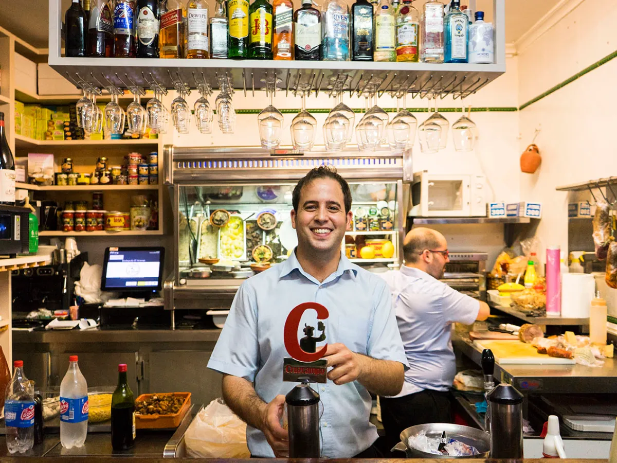 Man smiling and holding a red object in a bar with bottles and glasses overhead.
