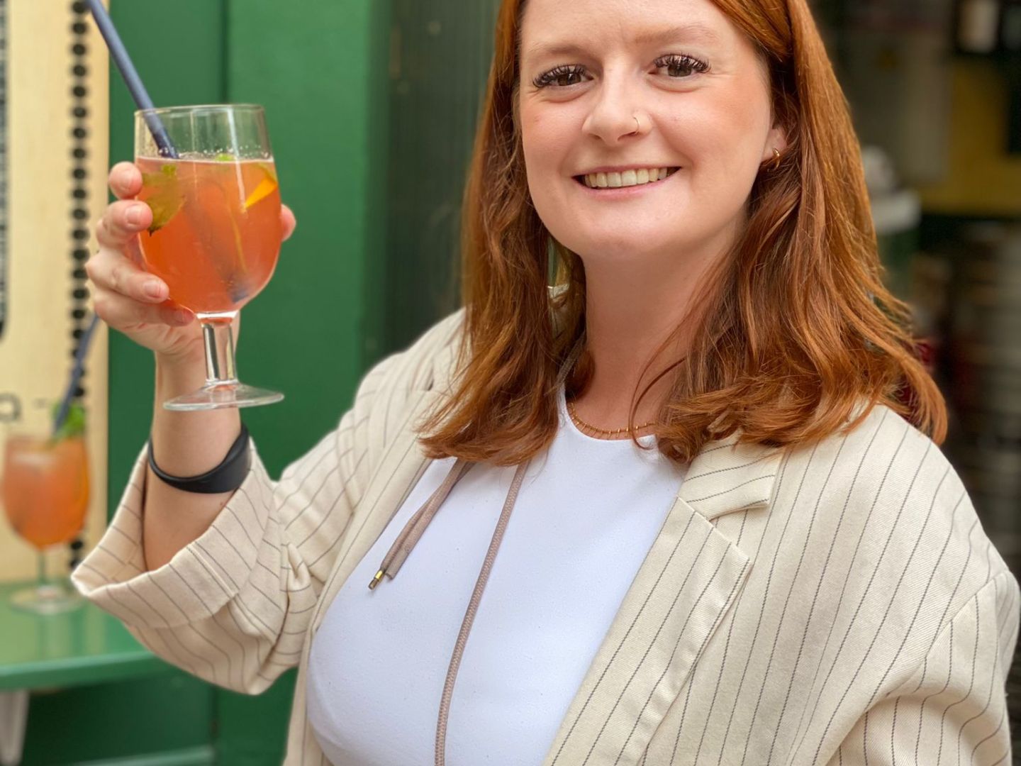 Woman holding a drink and a wooden platter with food, smiling outside a green building.