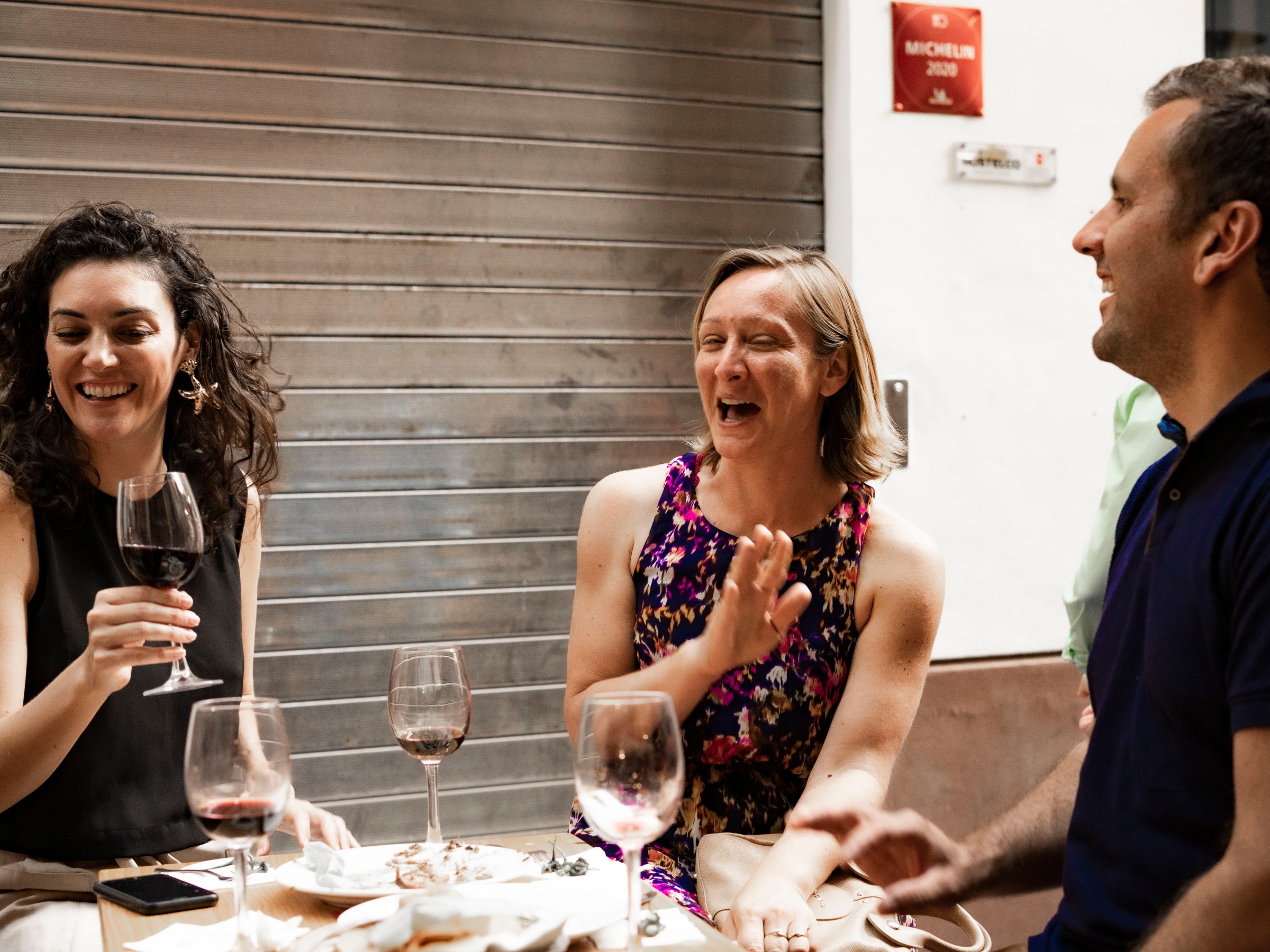 Three people laughing and enjoying wine at an outdoor table.