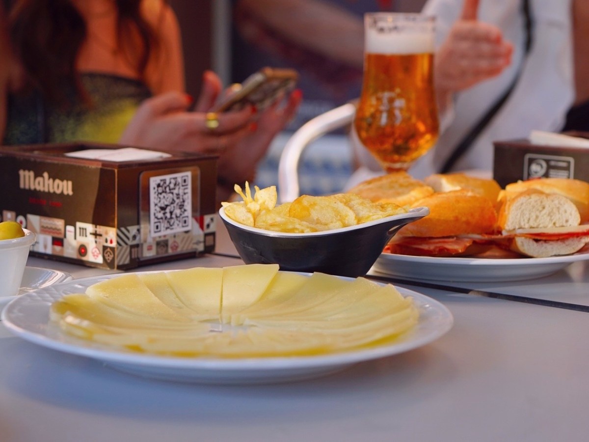 Table with cheese, chips, bread rolls, and olives; people in background, focus on food.
