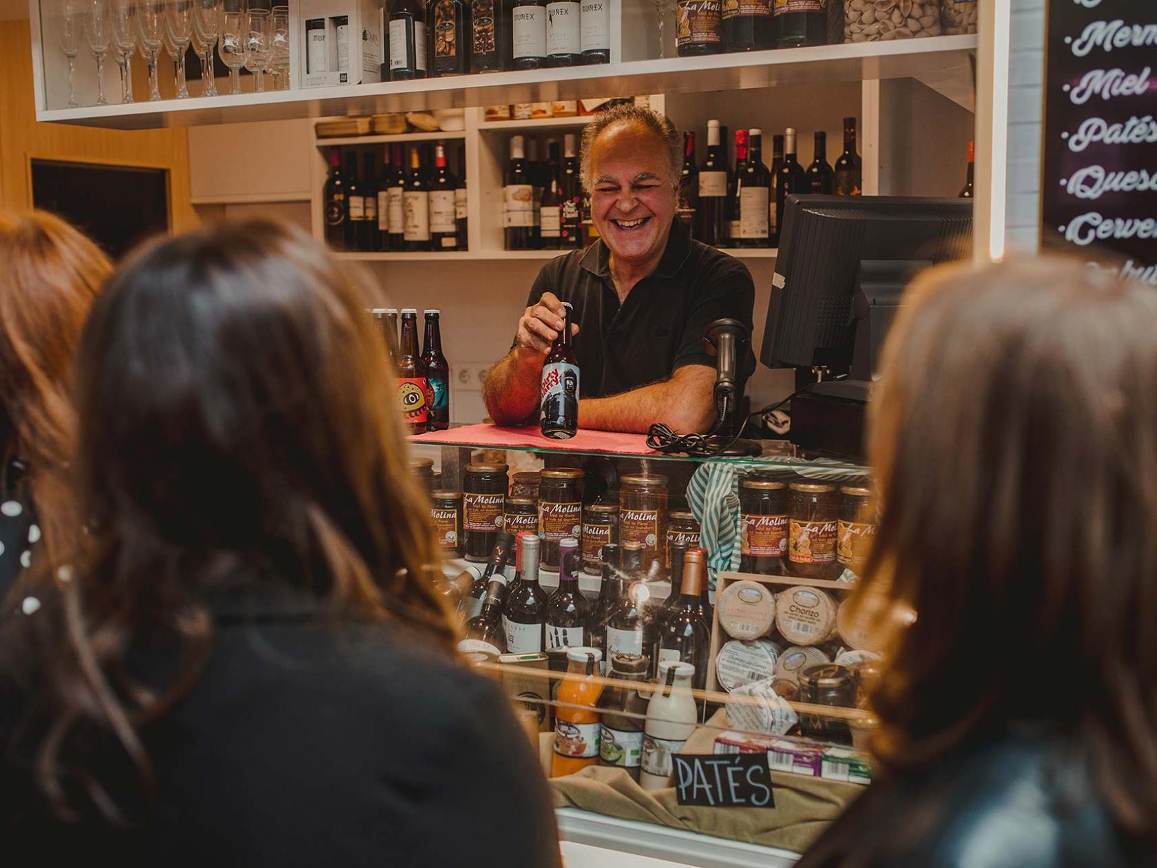 Smiling man behind deli counter with wine and jars, customers in foreground.