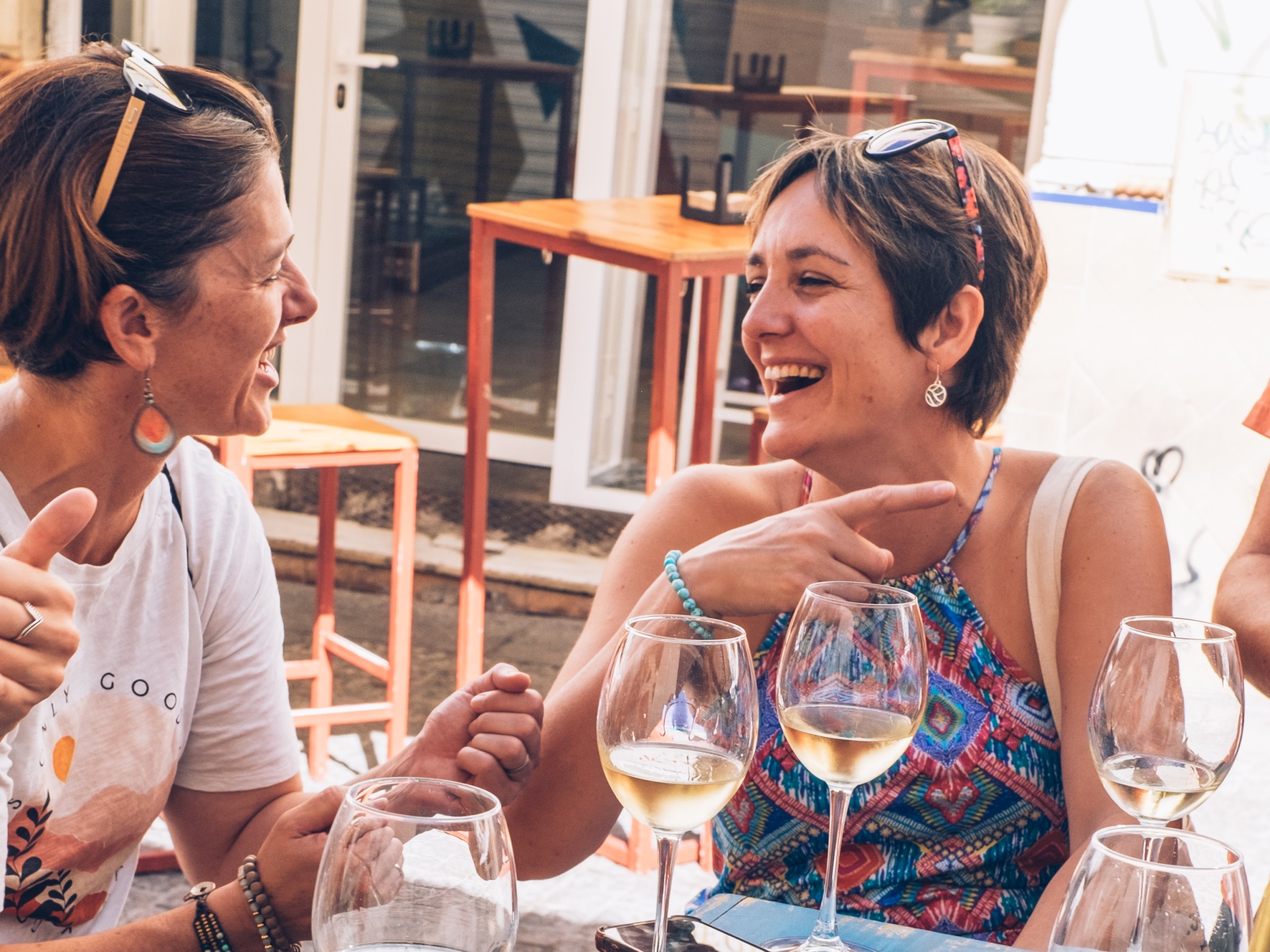 Two women laughing and talking at an outdoor café with wine glasses on the table.