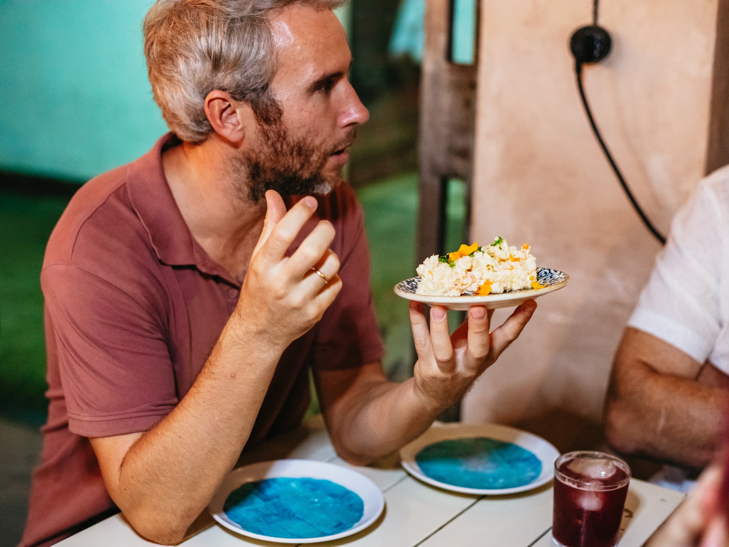 Man in a brown shirt holds a plate of food while talking at a table.