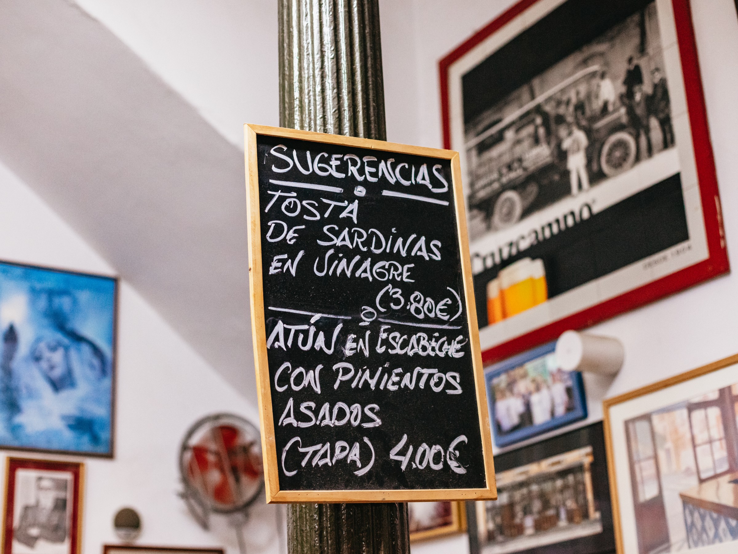 Chalkboard menu with Spanish food items, framed photos on wall in background.