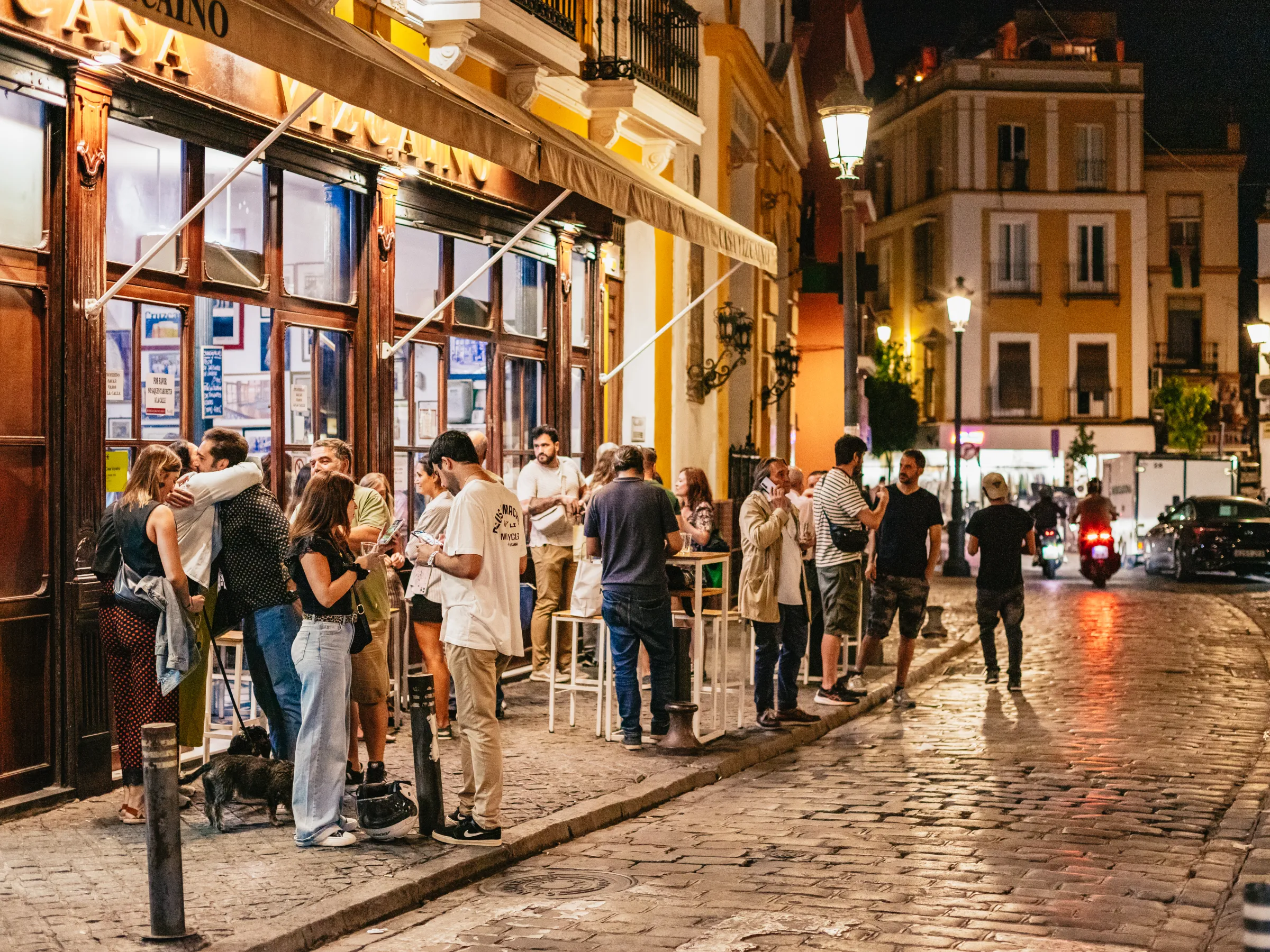 People socializing outside a bar on a lively, cobblestone street at night.