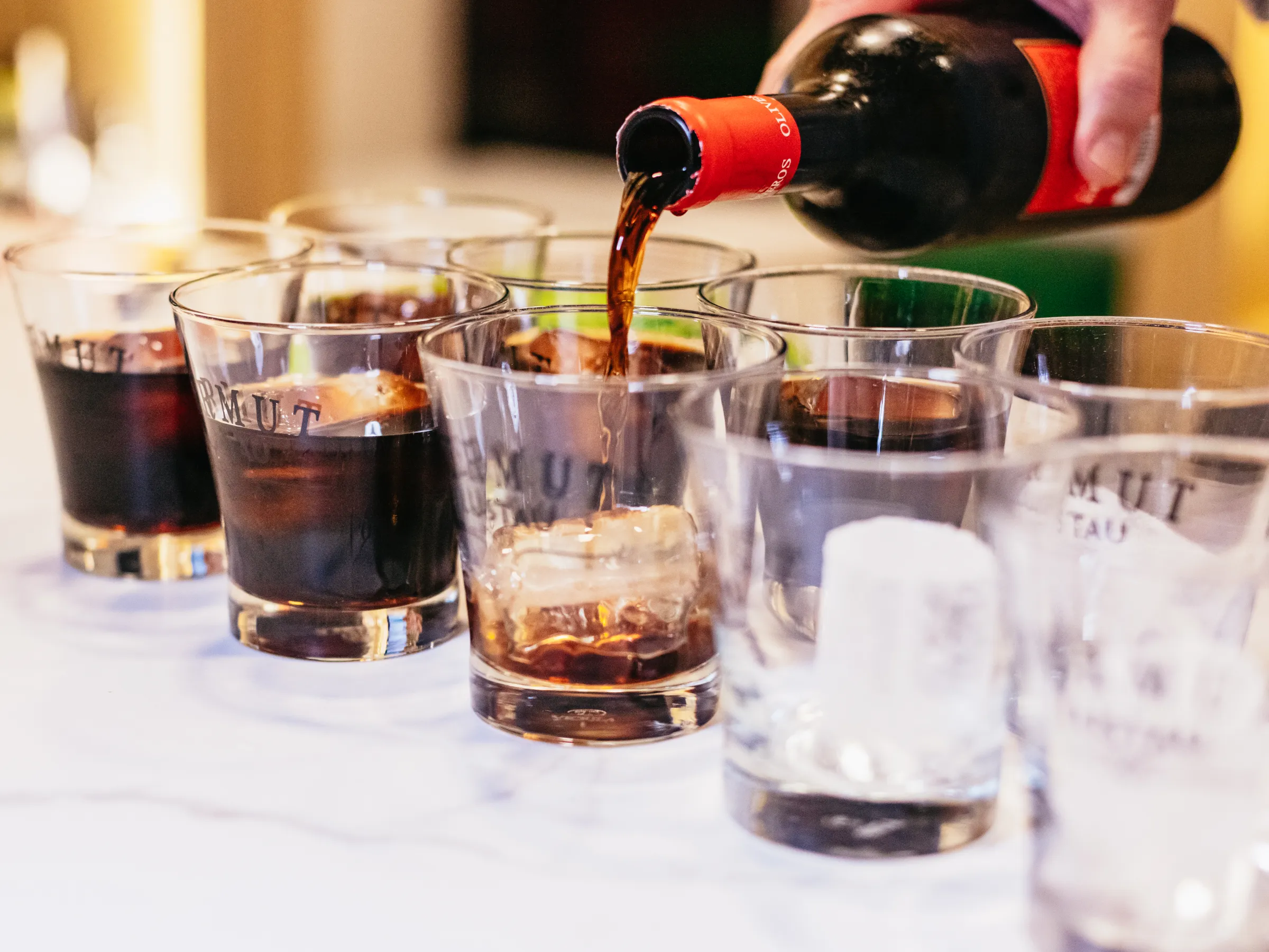 Hand pouring dark beverage into glasses with ice on a table.