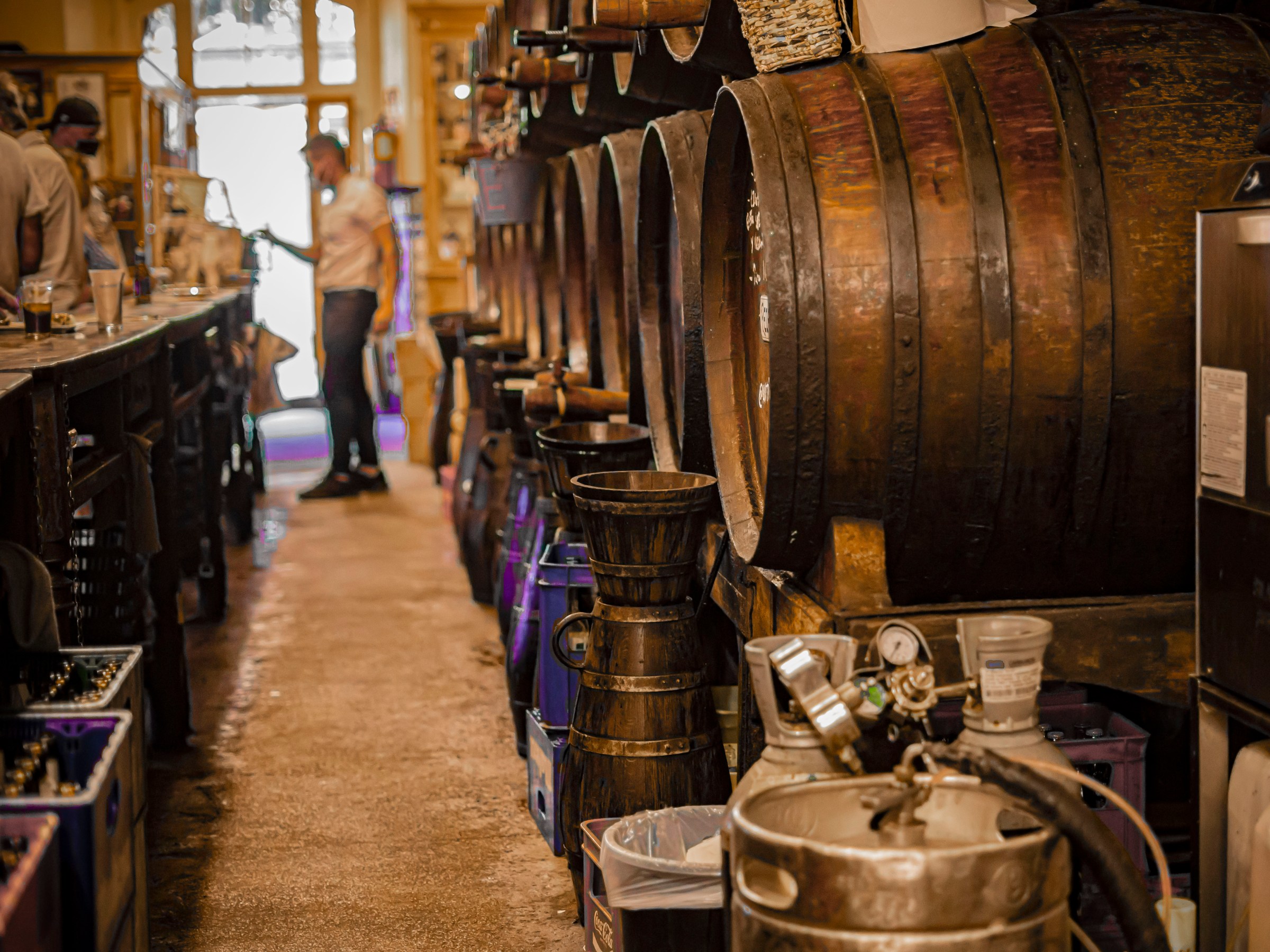 Bar interior with large wooden barrels on right and people seated at counter on left.
