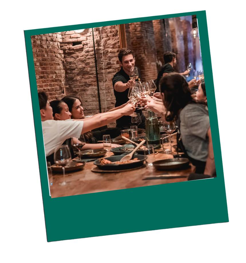 Group of people toasting with wine glasses around a wooden dining table in a brick-walled room.