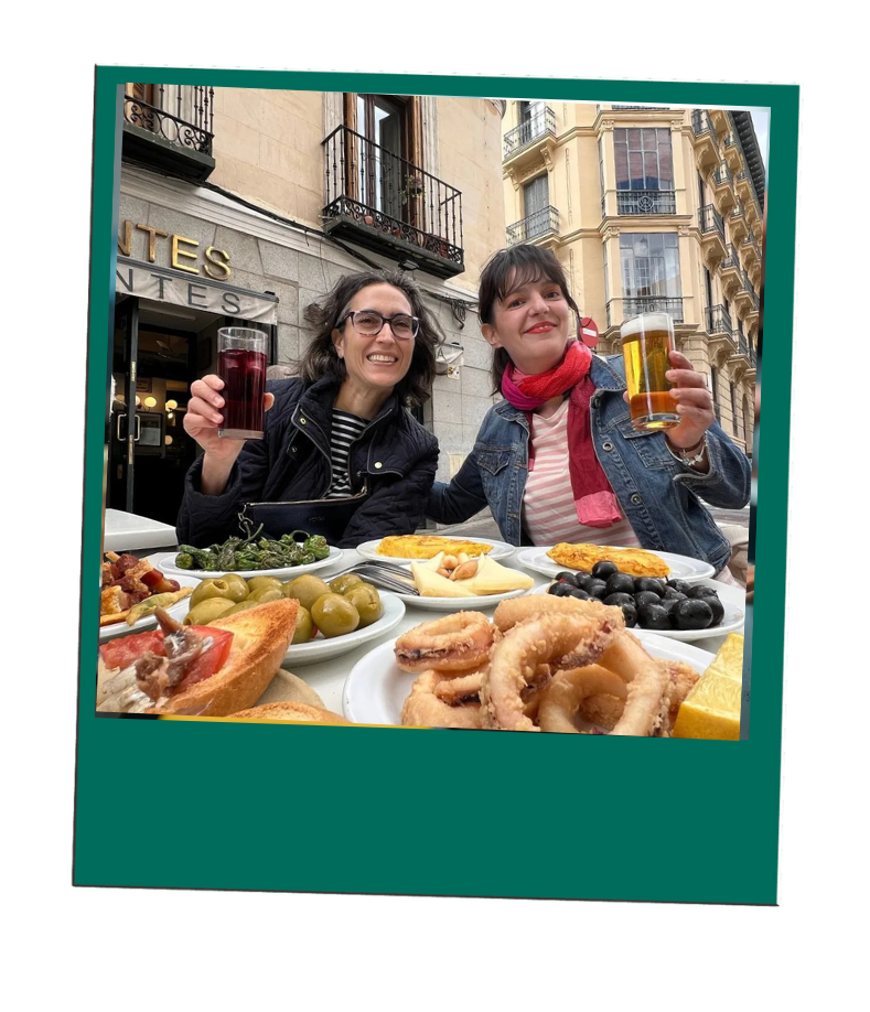 Two women toast with drinks at a table full of tapas in an outdoor café.