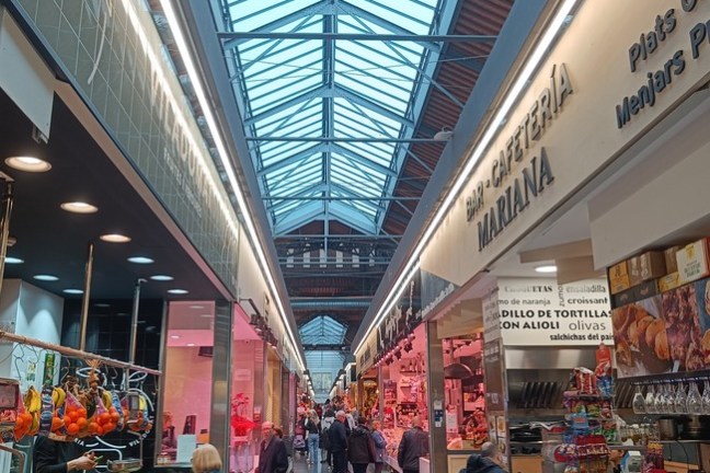 Indoor market with glass roof, shops on both sides, and people walking through the aisle.