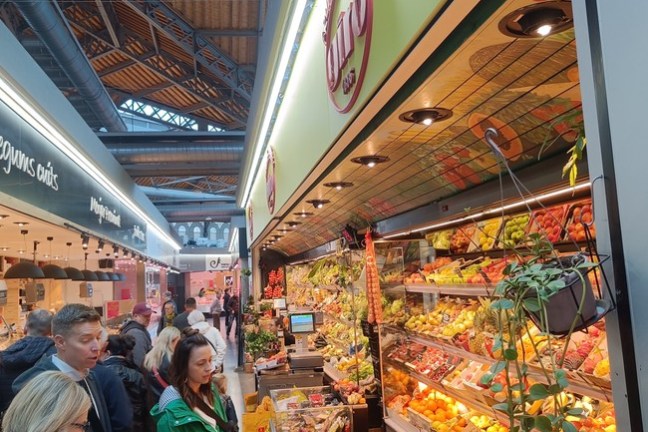 People shopping at a colorful indoor fruit and vegetable market stall.