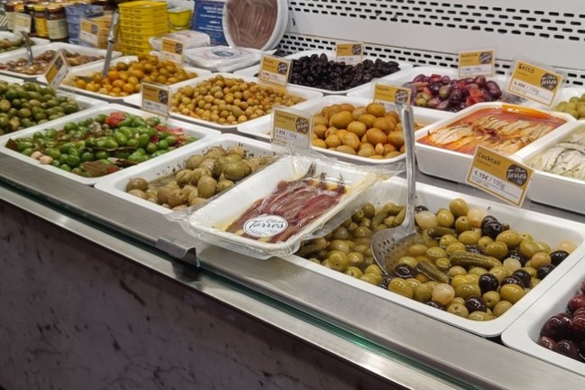 Market display of assorted olives and pickled items in trays with price tags.