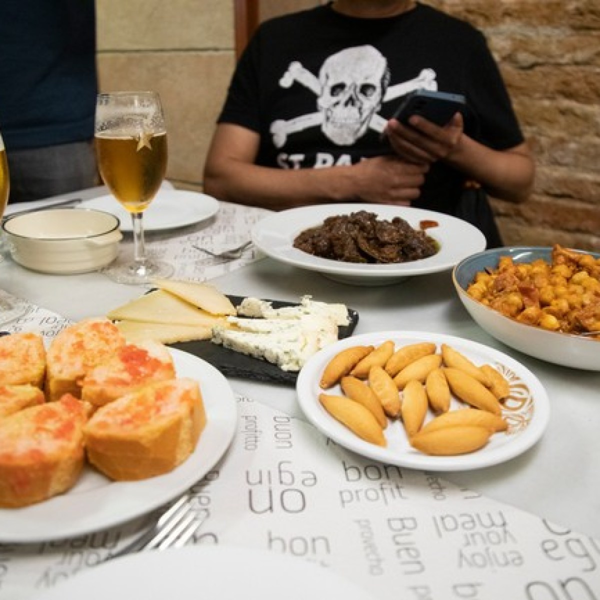 Table with tapas, cheese, meats, bread, and two glasses of beer.