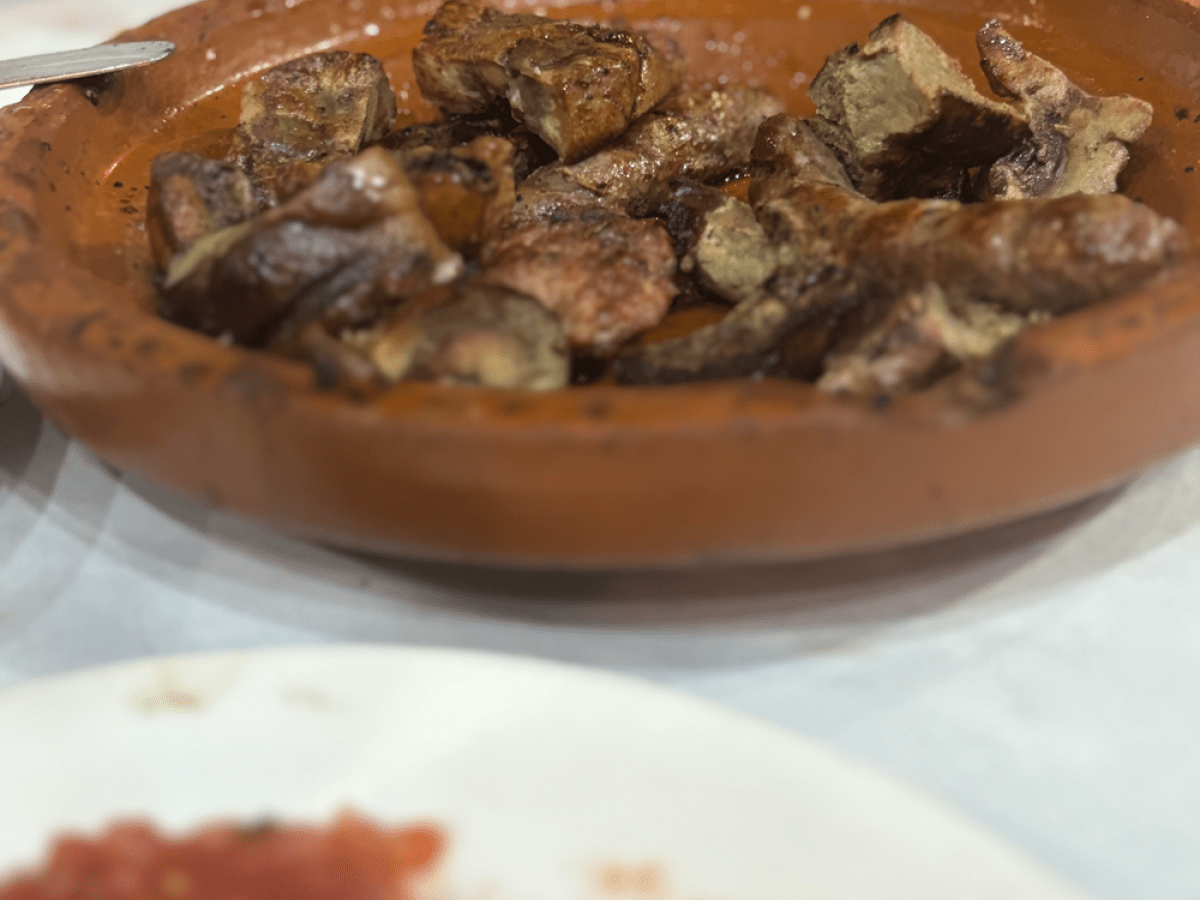 Clay dish with cooked meat pieces on a table near a plate of tomato sauce.