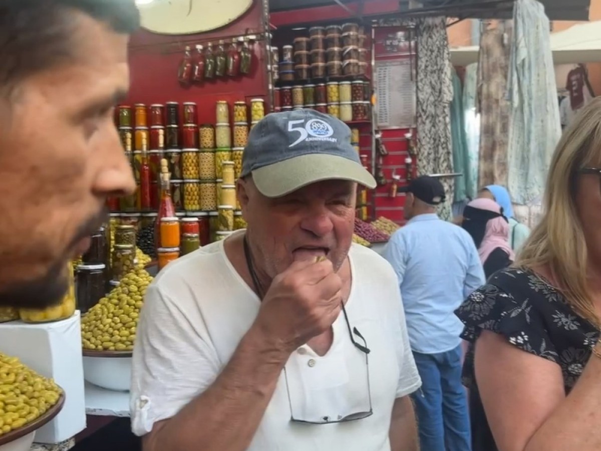 People tasting olives at an outdoor market stall filled with jars and baskets of produce.